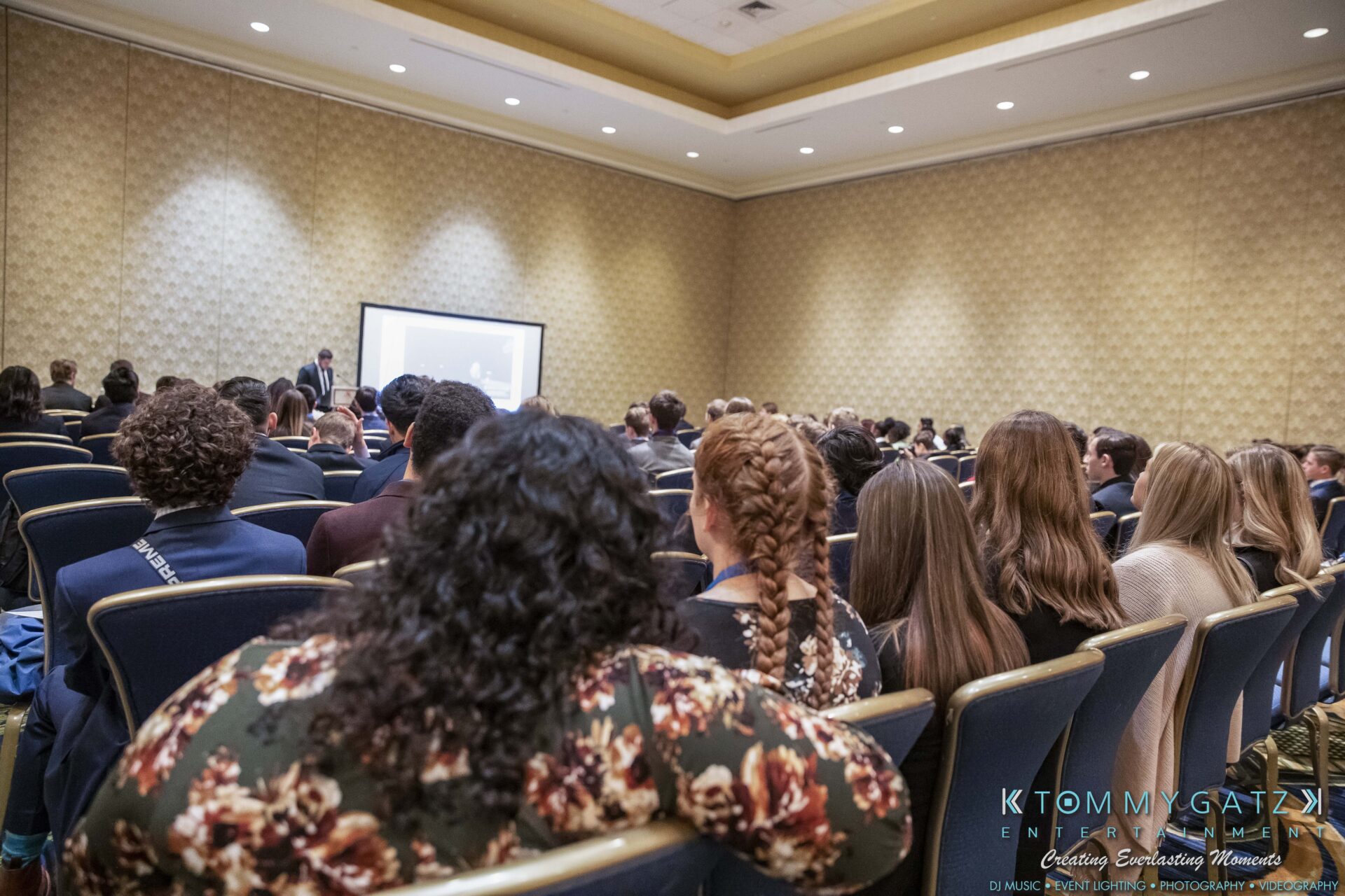 DECA-2018-Low-Res-WM-9 - TOMMY GATZ ENTERTAINMENT Audience attentively watching a presentation in a conference room with patterned walls and rows of chairs.