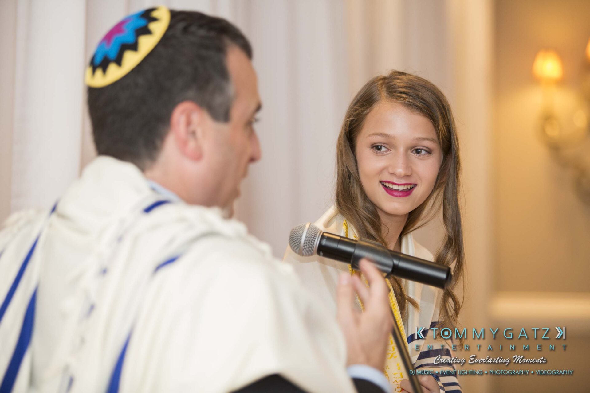Dylans-Bat-Mitzvah-CEREMONY-Low-Res-WM-121 - TOMMY GATZ ENTERTAINMENT A person in a prayer shawl and yarmulke speaks into a microphone while facing a smiling young girl.