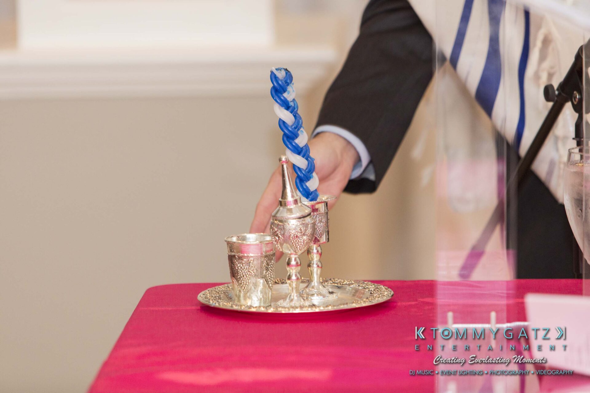 Dylans-Bat-Mitzvah-CEREMONY-Low-Res-WM-129 - TOMMY GATZ ENTERTAINMENT A silver havdalah set, including a braided candle, spice box, and wine cup, displayed on a table with a pink cloth.
