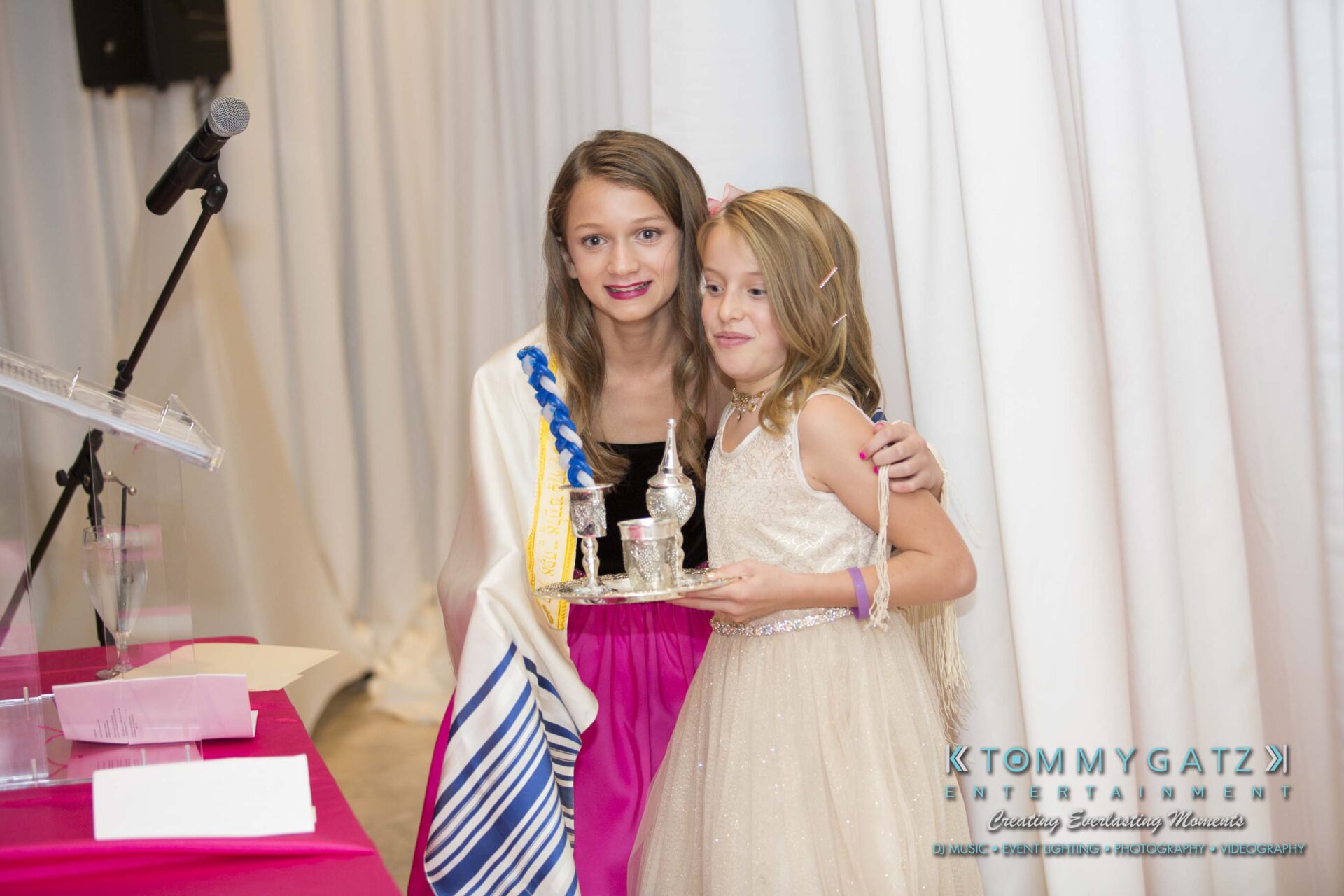 Dylans-Bat-Mitzvah-CEREMONY-Low-Res-WM-134 - TOMMY GATZ ENTERTAINMENT Two children, one holding a small trophy, are standing indoors at an event. A podium and microphone are visible nearby.