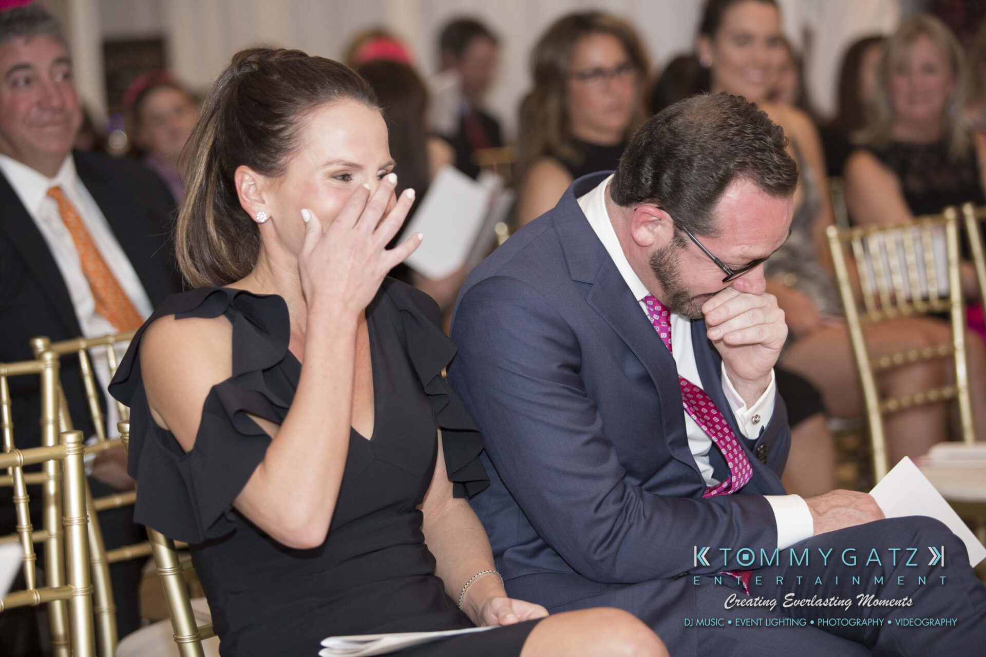 Dylans-Bat-Mitzvah-CEREMONY-Low-Res-WM-144 - TOMMY GATZ ENTERTAINMENT A woman and a man sitting on chairs, both appear to be laughing. The man is covering his mouth with a paper. Other people are seated in the background.