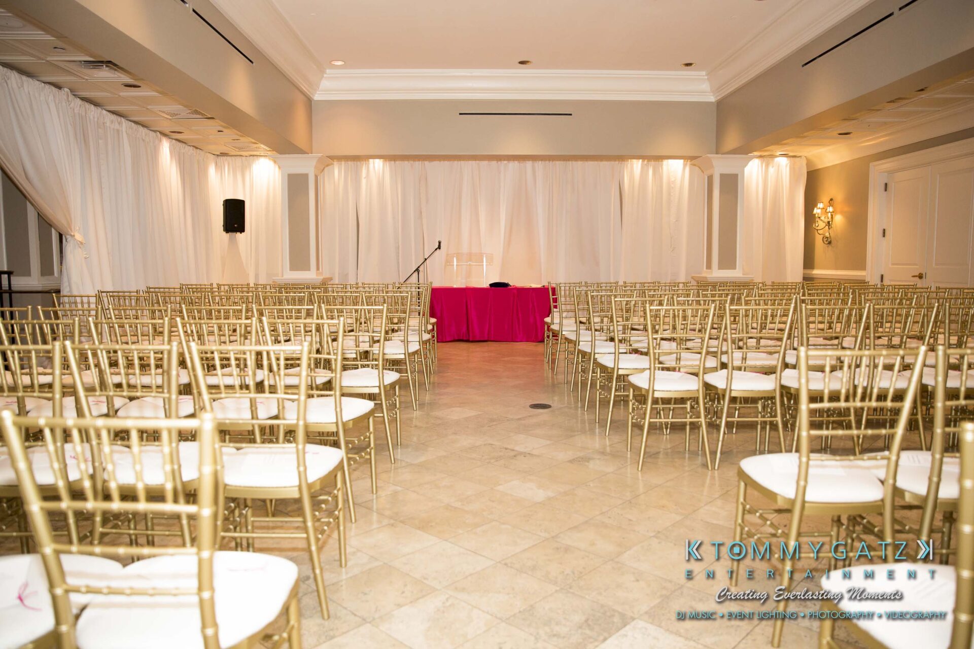 Dylans-Bat-Mitzvah-CEREMONY-Low-Res-WM-15 - TOMMY GATZ ENTERTAINMENT An empty event venue with rows of gold chairs facing a stage with pink tablecloth and a microphone stand. White drapes cover part of the walls.