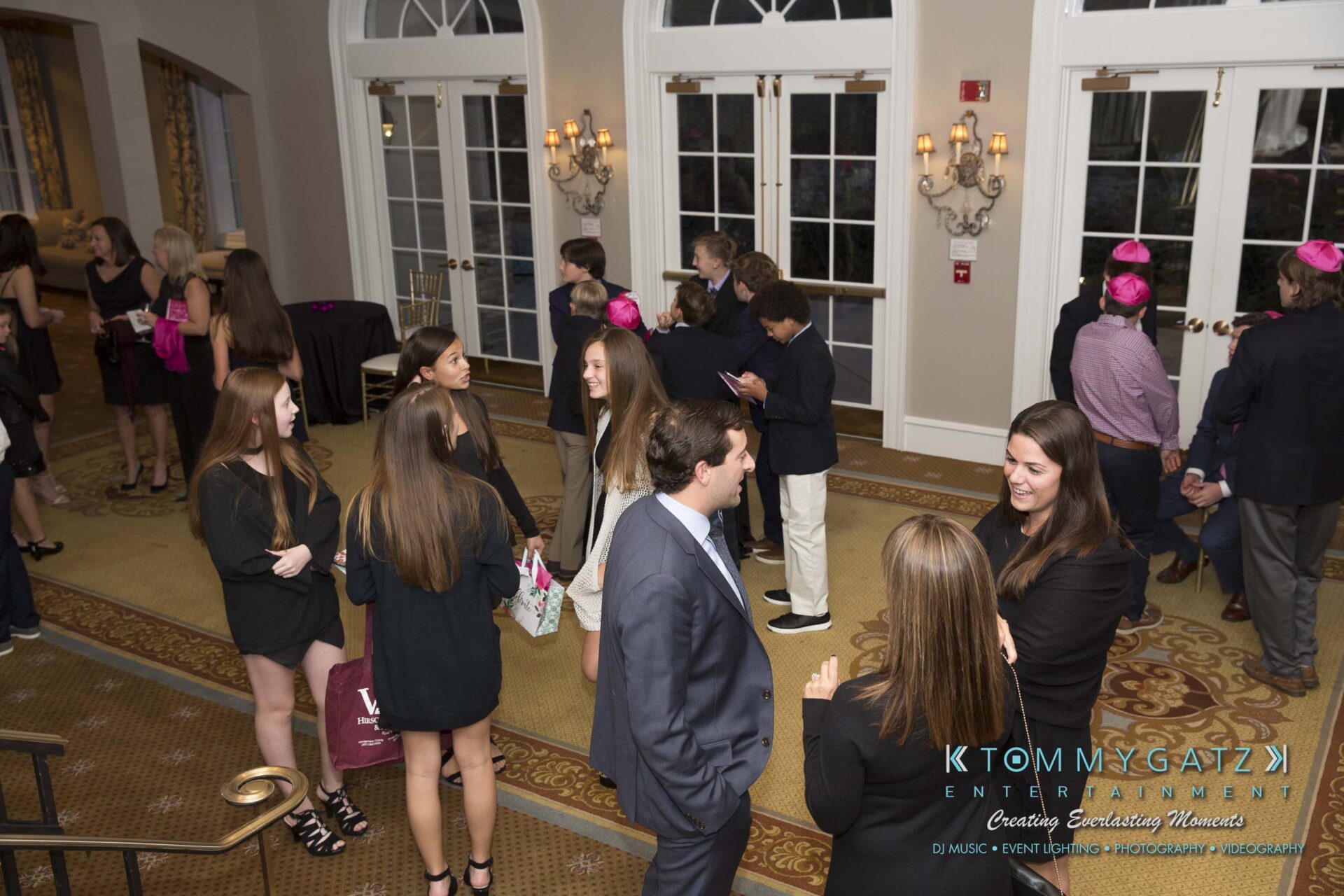 Dylans-Bat-Mitzvah-CEREMONY-Low-Res-WM-18 - TOMMY GATZ ENTERTAINMENT A group of people socializing in a banquet hall with large windows and carpeting. Some are wearing pink hats. A staircase is visible on the left.