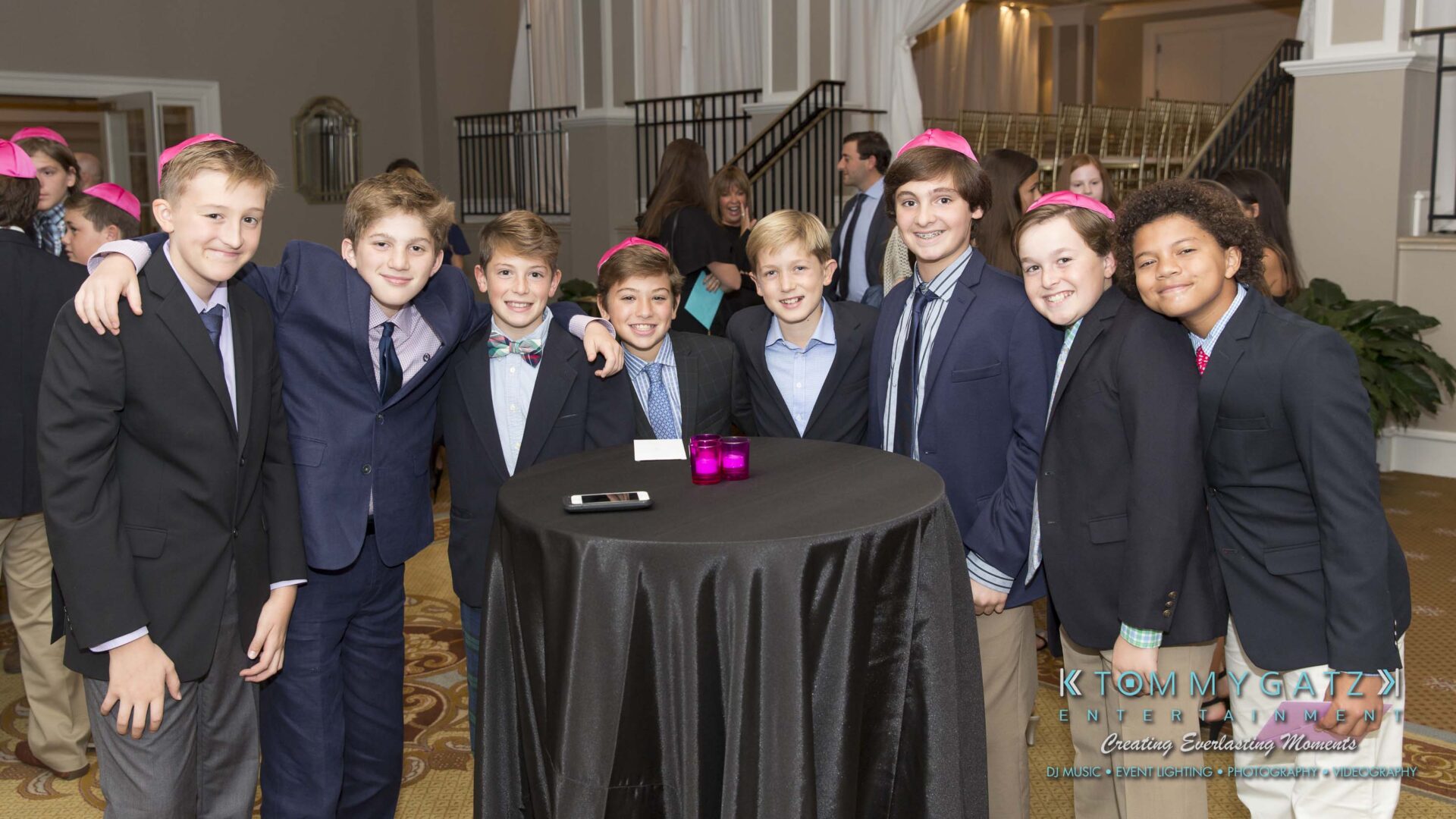 Dylans-Bat-Mitzvah-CEREMONY-Low-Res-WM-20 - TOMMY GATZ ENTERTAINMENT Group of boys in suits and pink yarmulkes smile around a tall table at a formal event.