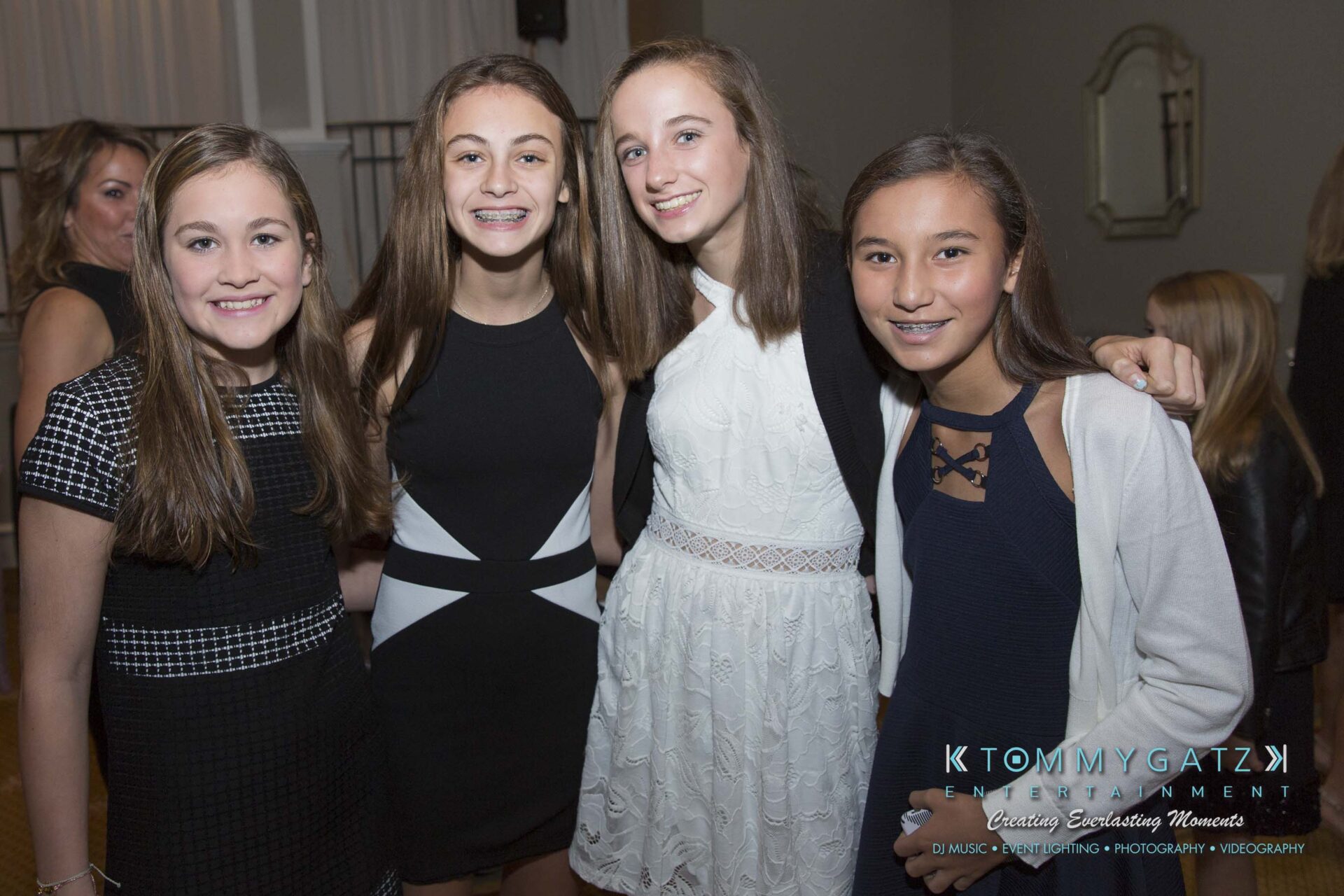 Dylans-Bat-Mitzvah-CEREMONY-Low-Res-WM-23 - TOMMY GATZ ENTERTAINMENT Four young women smiling at the camera, dressed in formal attire at an indoor event.