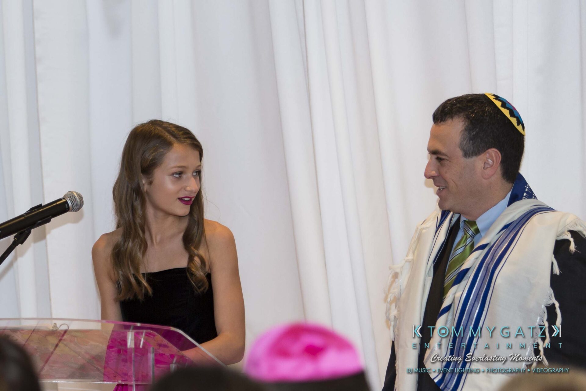 Dylans-Bat-Mitzvah-CEREMONY-Low-Res-WM-54 - TOMMY GATZ ENTERTAINMENT A young girl and a man wearing a tallit are standing at a podium. The girl looks at the man as he appears to speak. A microphone is positioned near them.