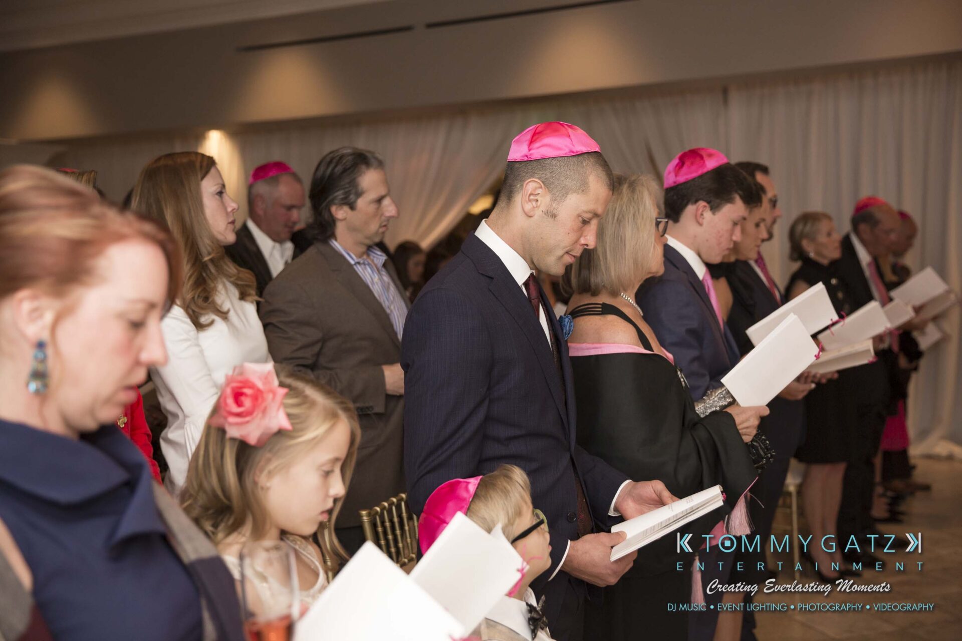 Dylans-Bat-Mitzvah-CEREMONY-Low-Res-WM-58 - TOMMY GATZ ENTERTAINMENT A group of people in formal attire are standing and holding booklets. Many are wearing pink caps.