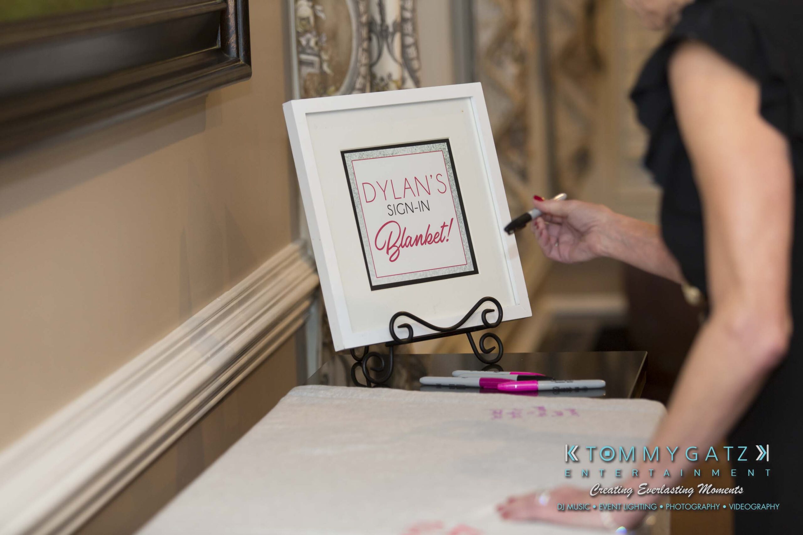 Dylans-Bat-Mitzvah-CEREMONY-Low-Res-WM-6 - TOMMY GATZ ENTERTAINMENT A person writes on a tablecloth next to a framed sign reading "Dylan's Sign-In Blanket" with markers on the table.