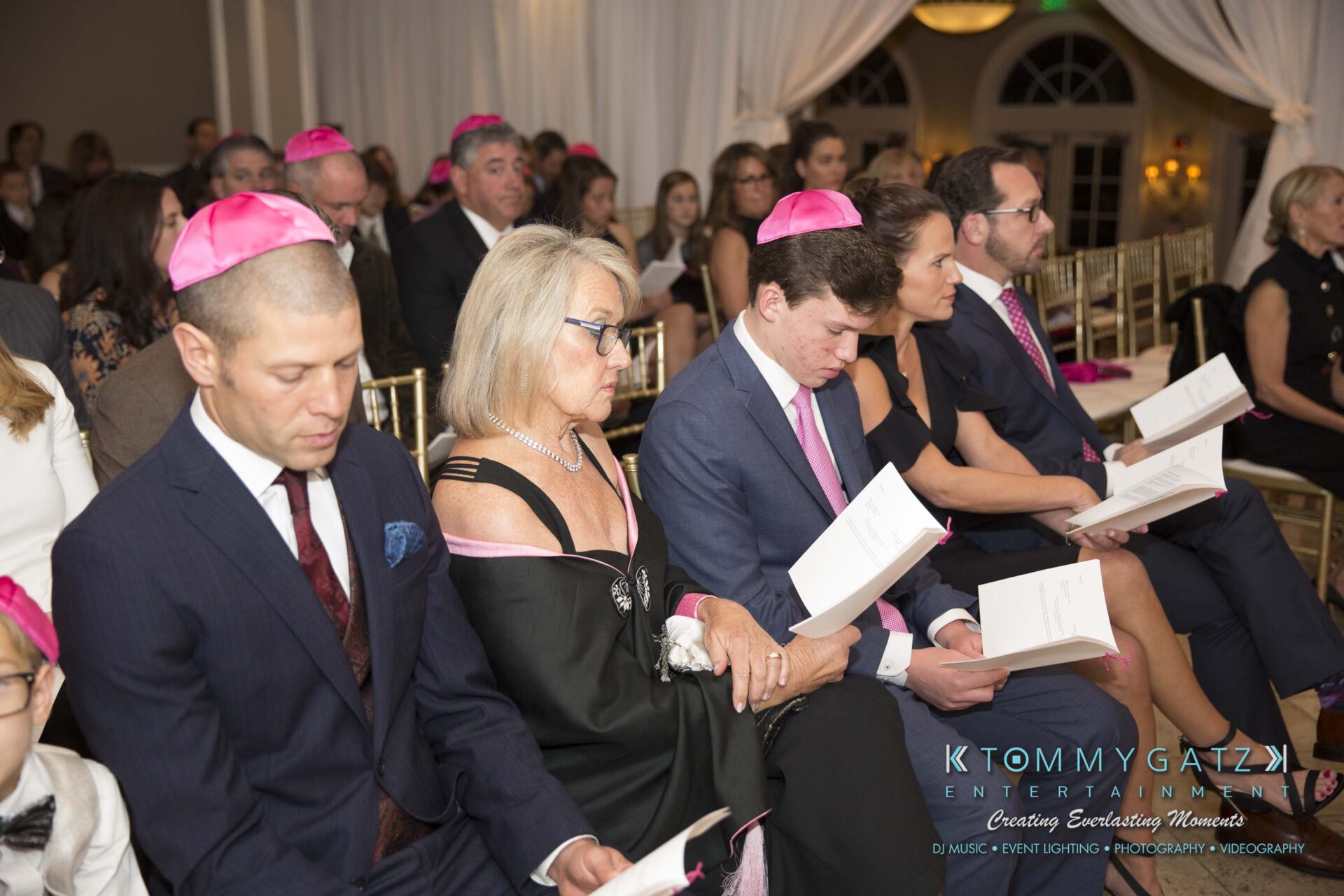 Dylans-Bat-Mitzvah-CEREMONY-Low-Res-WM-63 - TOMMY GATZ ENTERTAINMENT People seated in a formal event, wearing pink yarmulkes and holding papers, attentive to proceedings.