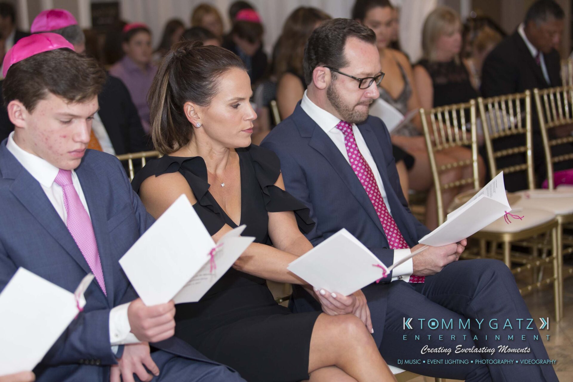 Dylans-Bat-Mitzvah-CEREMONY-Low-Res-WM-67 - TOMMY GATZ ENTERTAINMENT Three people sit in a row of gold chairs, wearing formal attire, reading booklets.