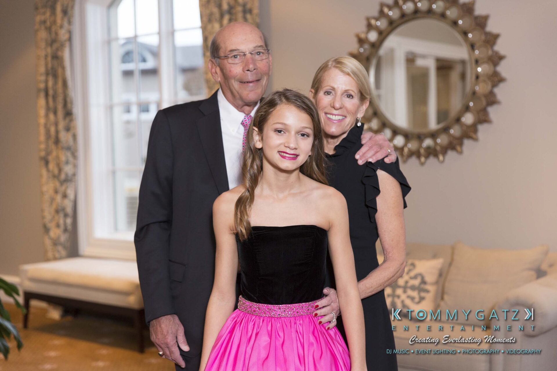 Dylans-Bat-Mitzvah-PORTRAITS-Low-Res-WM-35 - TOMMY GATZ ENTERTAINMENT A young girl in a pink dress poses with an older man and woman inside a room with a mirror and two windows.