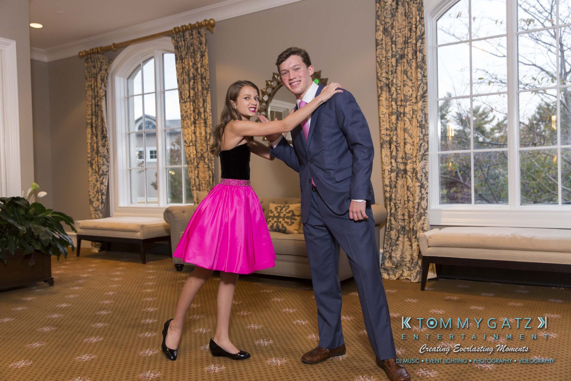Dylans-Bat-Mitzvah-PORTRAITS-Low-Res-WM-36 - TOMMY GATZ ENTERTAINMENT A young woman in a pink dress playfully poses with a young man in a suit in a room with large windows and beige carpet.