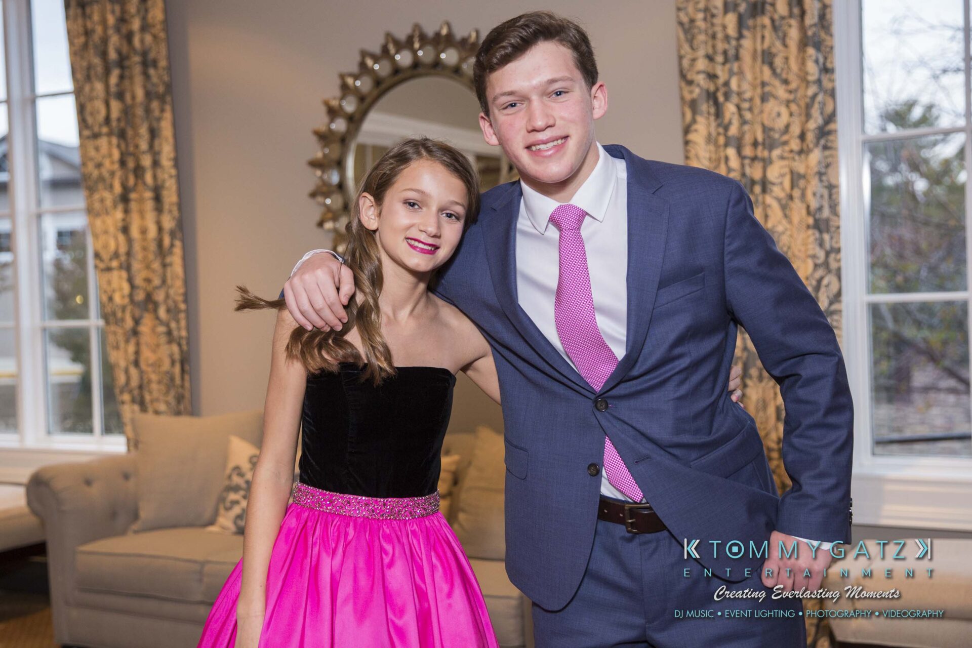 Dylans-Bat-Mitzvah-PORTRAITS-Low-Res-WM-39 - TOMMY GATZ ENTERTAINMENT A young girl and boy, both dressed in formal wear, pose together in a living room. The girl wears a pink dress, and the boy wears a suit with a pink tie.