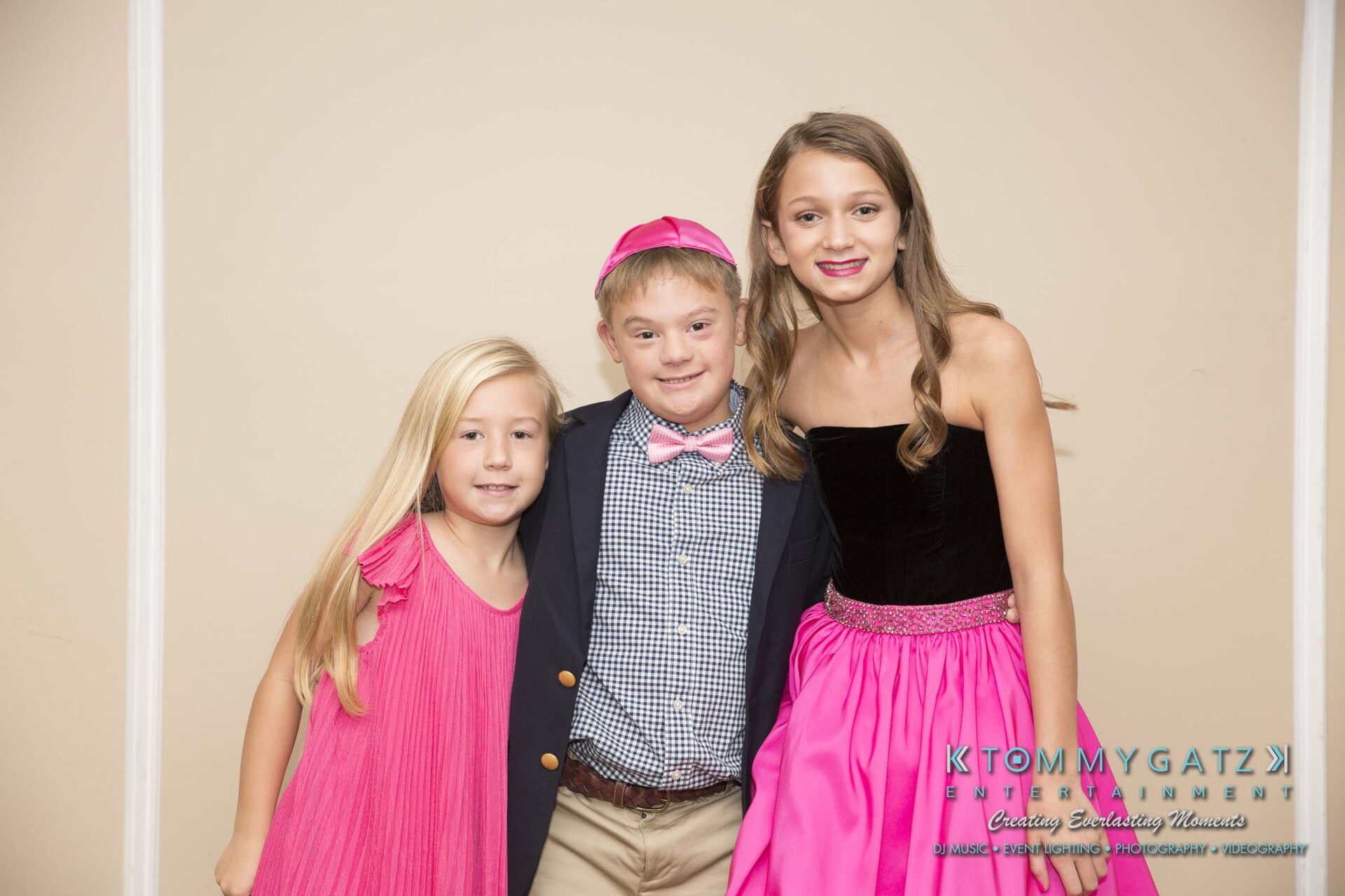 Dylans-Bat-Mitzvah-PORTRAITS-Low-Res-WM-60 - TOMMY GATZ ENTERTAINMENT Three children in pink outfits stand together, smiling in front of a beige wall. The boy in the center wears a pink bow tie and yarmulke.