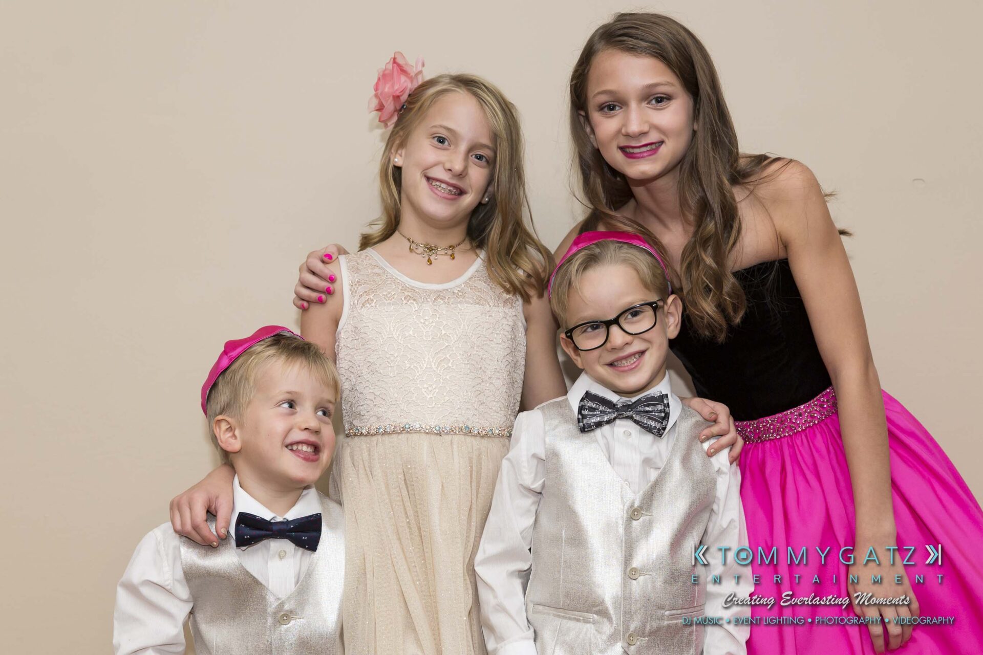 Dylans-Bat-Mitzvah-PORTRAITS-Low-Res-WM-65 - TOMMY GATZ ENTERTAINMENT Four children dressed in formal attire smiling and posing together against a plain background. Two girls in dresses, two boys in vests and bow ties.
