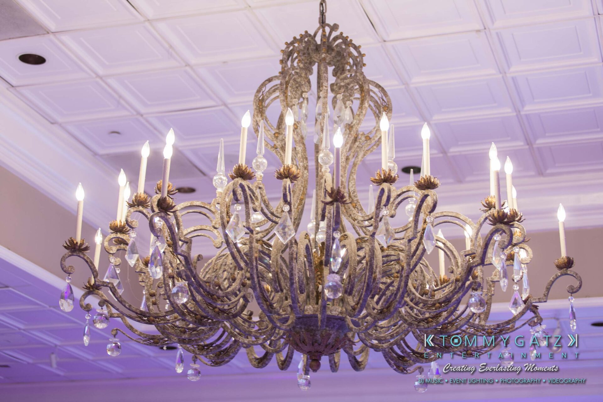 Dylans-Bat-Mitzvah-RECEPTION-Low-Res-WM-1 - TOMMY GATZ ENTERTAINMENT An ornate chandelier with lit candles hangs from a white-paneled ceiling.