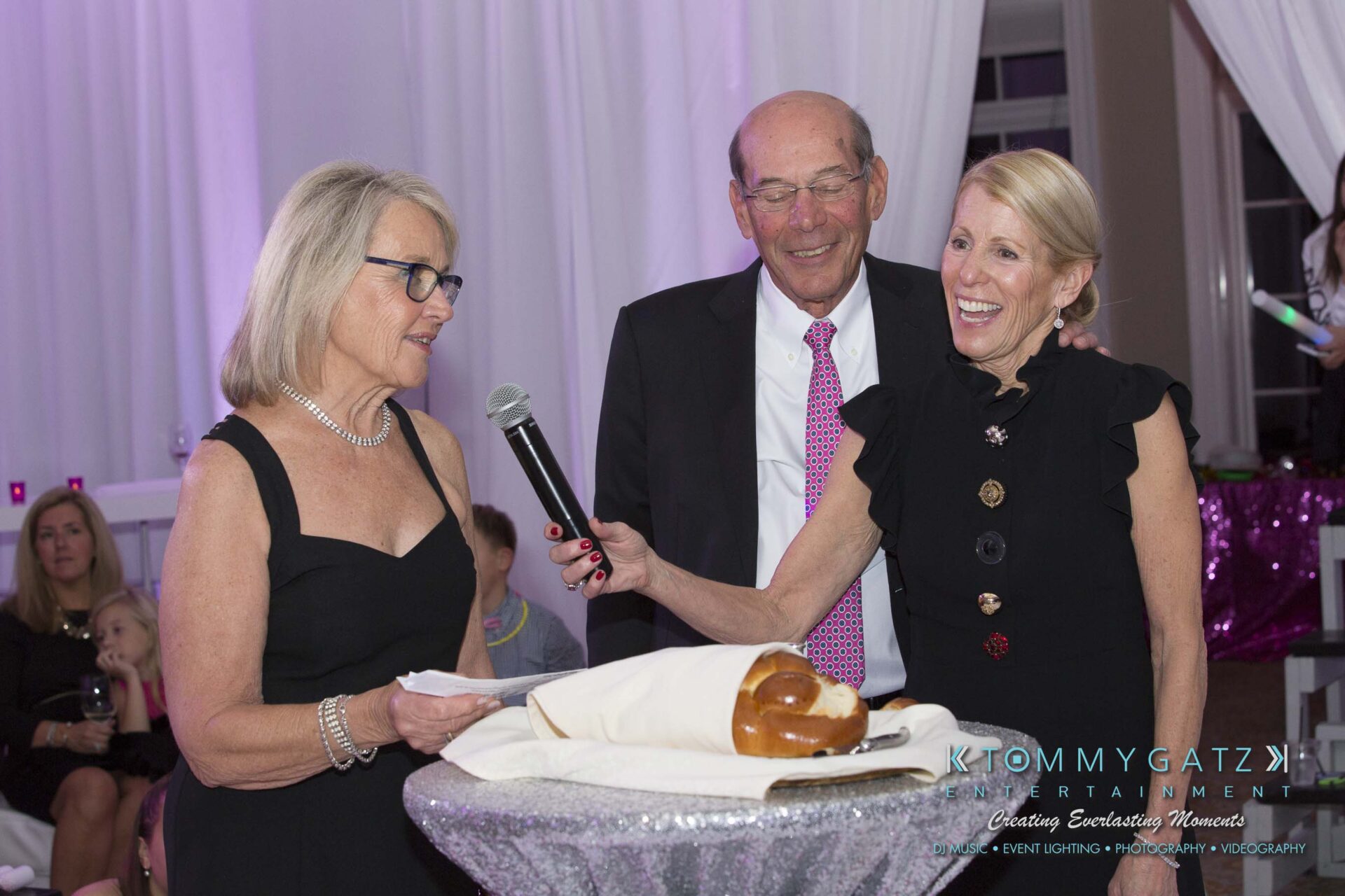 Dylans-Bat-Mitzvah-RECEPTION-Low-Res-WM-116 - TOMMY GATZ ENTERTAINMENT Three people stand together, one holding a microphone. A challah bread is on the table in front of them. The setting appears to be a celebratory event.