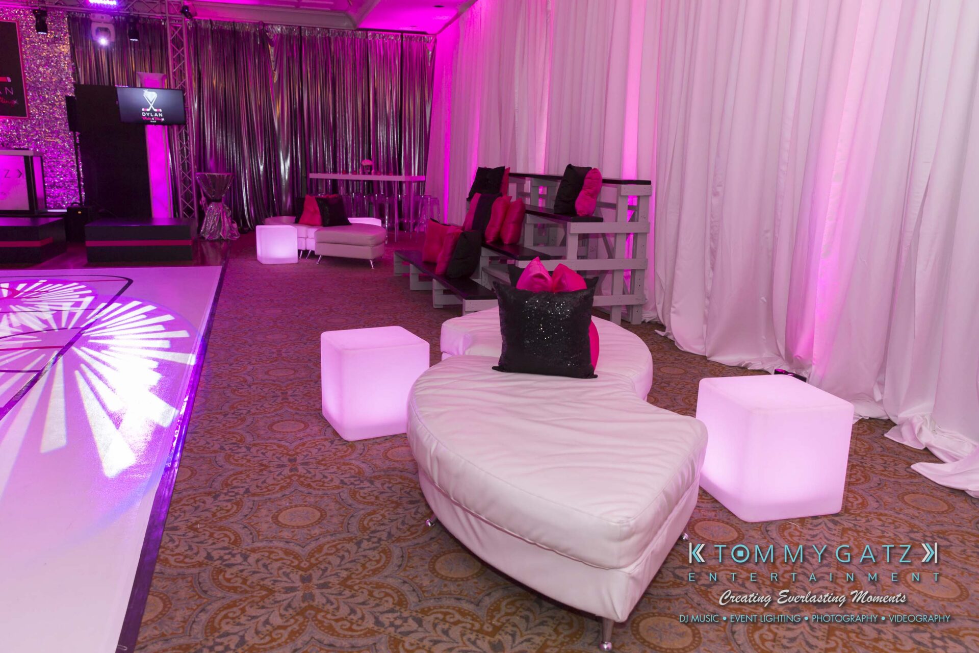 Dylans-Bat-Mitzvah-RECEPTION-Low-Res-WM-12 - TOMMY GATZ ENTERTAINMENT A lounge area with curved white seating, glowing cube tables, pink and black cushions, and pink lighting.