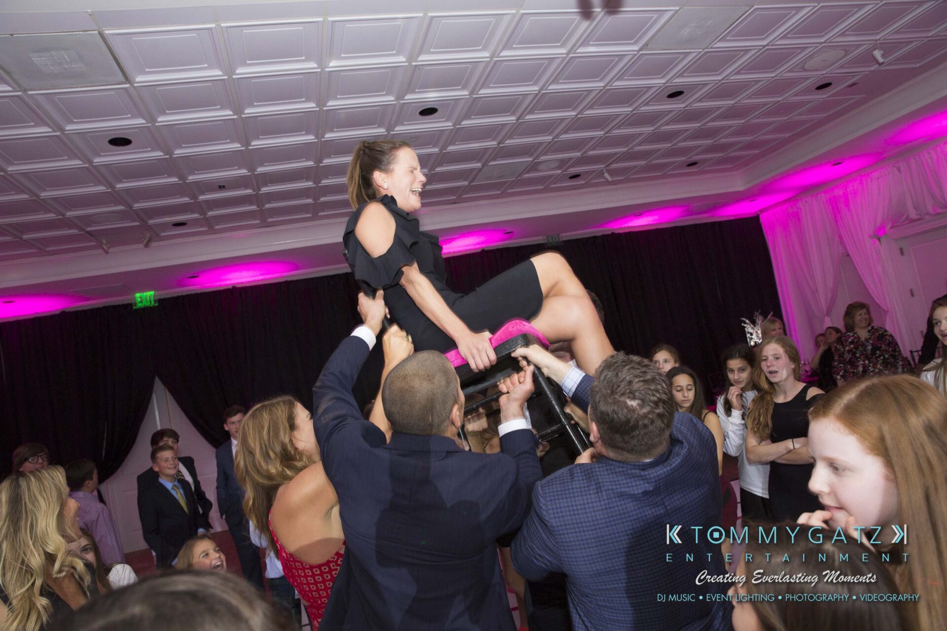Dylans-Bat-Mitzvah-RECEPTION-Low-Res-WM-153 - TOMMY GATZ ENTERTAINMENT Guests lift a woman on a chair at an indoor celebration with pink lighting.