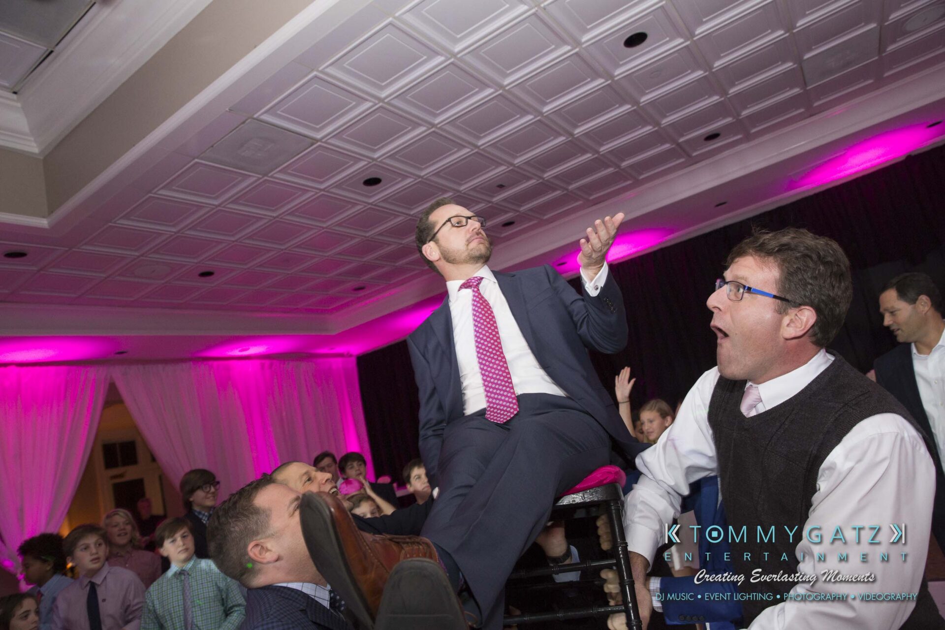 Dylans-Bat-Mitzvah-RECEPTION-Low-Res-WM-156 - TOMMY GATZ ENTERTAINMENT A man in a suit is lifted on a chair by two others at a lively indoor event. Pink and purple lighting fill the room where people watch and celebrate.
