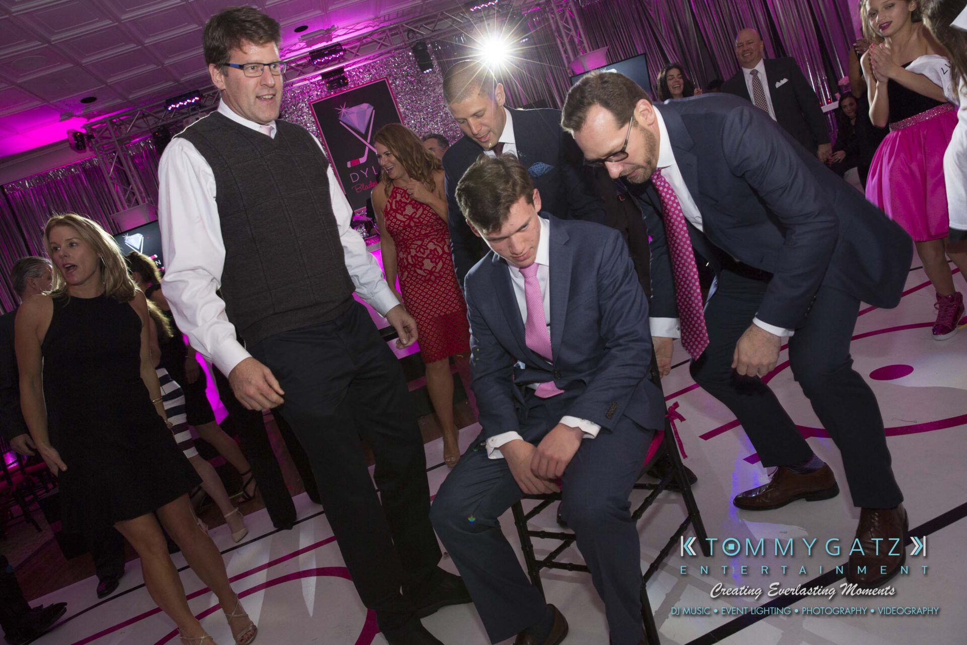 Dylans-Bat-Mitzvah-RECEPTION-Low-Res-WM-161 - TOMMY GATZ ENTERTAINMENT A group of people in formal attire gather around a seated person at an event with purple lighting.