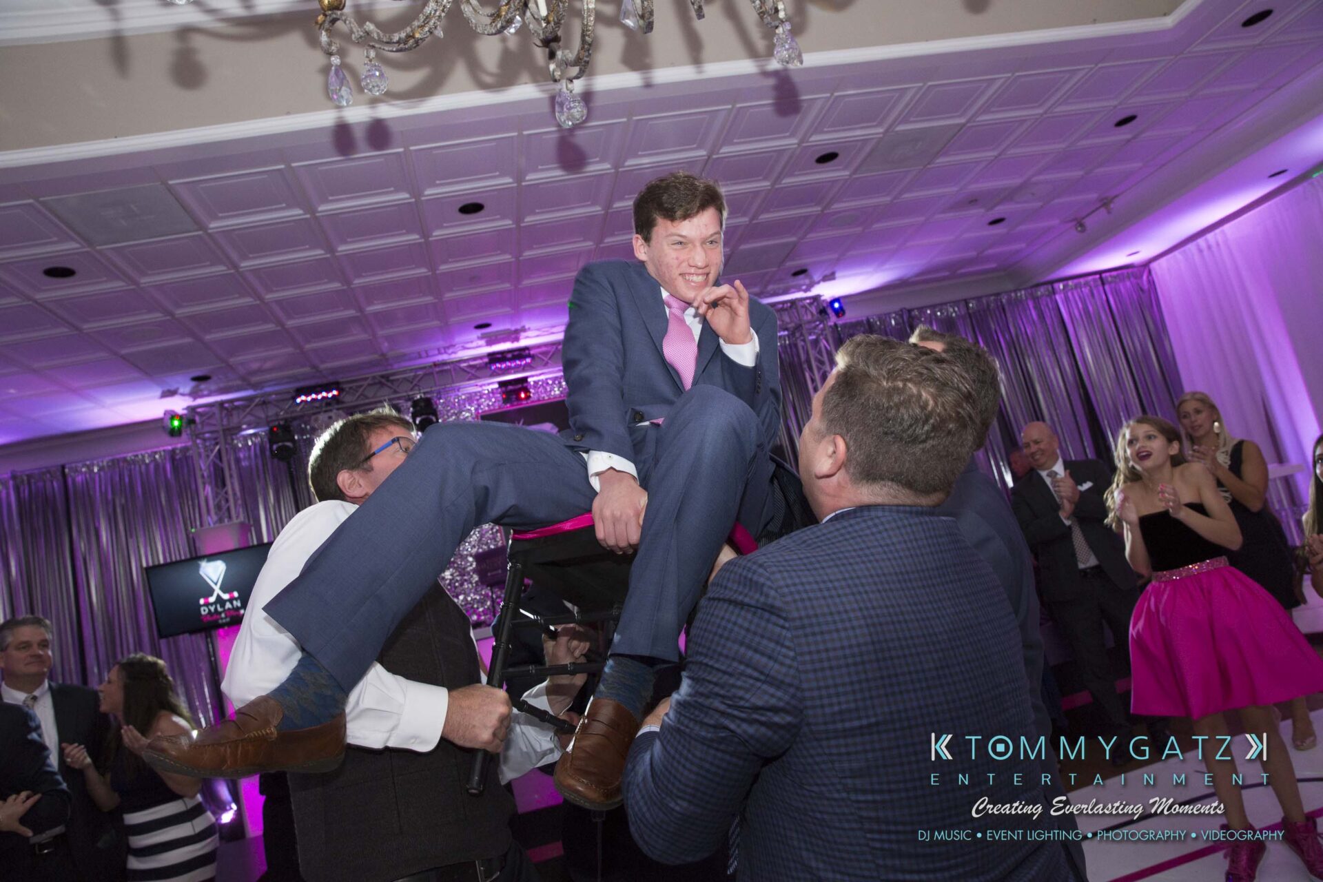 Dylans-Bat-Mitzvah-RECEPTION-Low-Res-WM-162 - TOMMY GATZ ENTERTAINMENT A man in a suit is lifted on a chair during a celebration in a ballroom, surrounded by people.