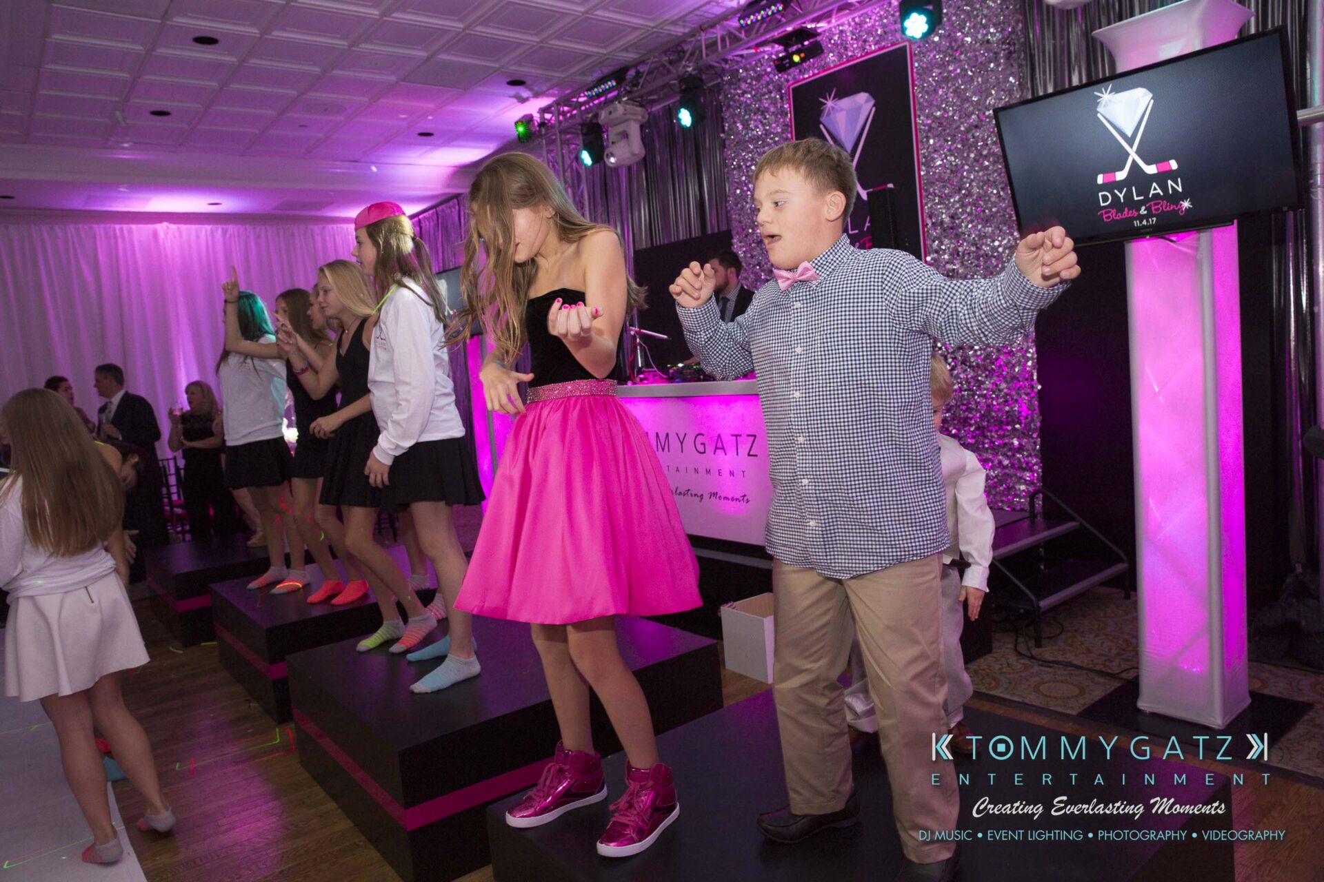Dylans-Bat-Mitzvah-RECEPTION-Low-Res-WM-170 - TOMMY GATZ ENTERTAINMENT Group of children dancing on a stage at a party, with pink and purple lights and a DJ booth in the background.