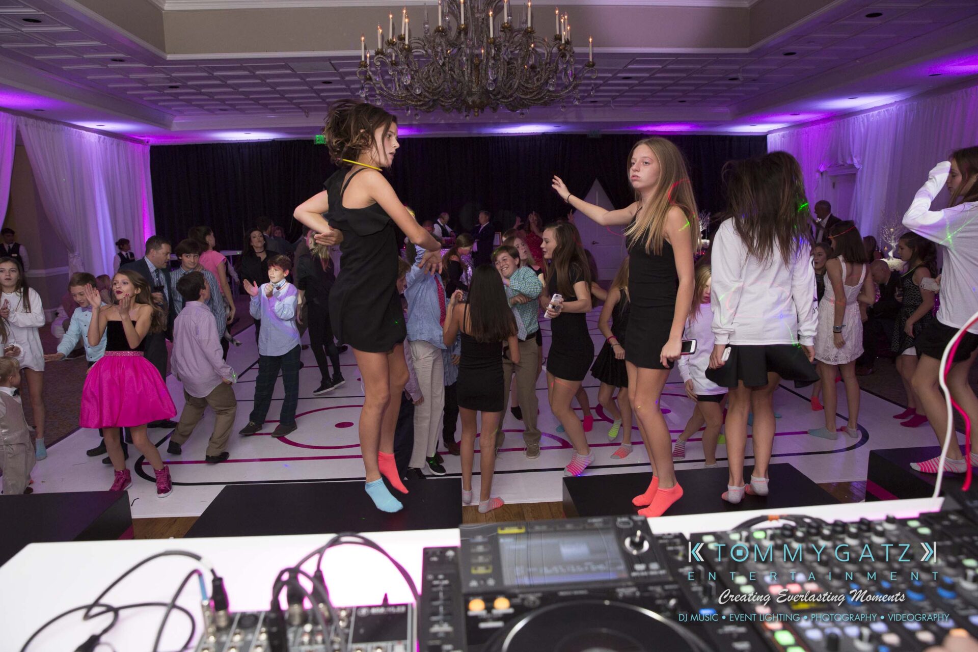 Dylans-Bat-Mitzvah-RECEPTION-Low-Res-WM-173 - TOMMY GATZ ENTERTAINMENT People dancing in a party venue with a DJ booth in the foreground and a chandelier overhead.