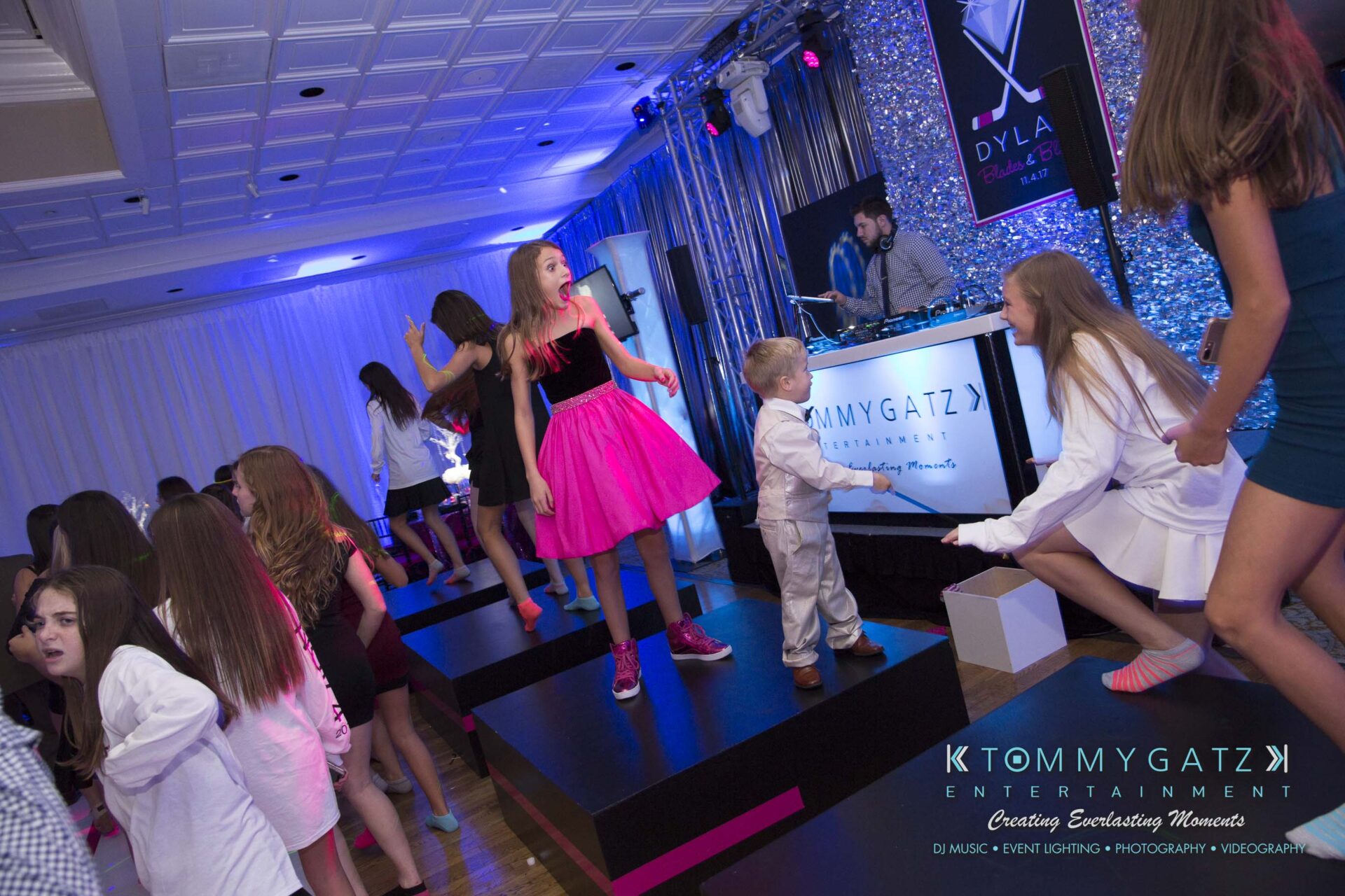 Dylans-Bat-Mitzvah-RECEPTION-Low-Res-WM-189 - TOMMY GATZ ENTERTAINMENT People dancing at a party with a DJ booth in the background. A girl in a pink dress stands out on the dance floor.