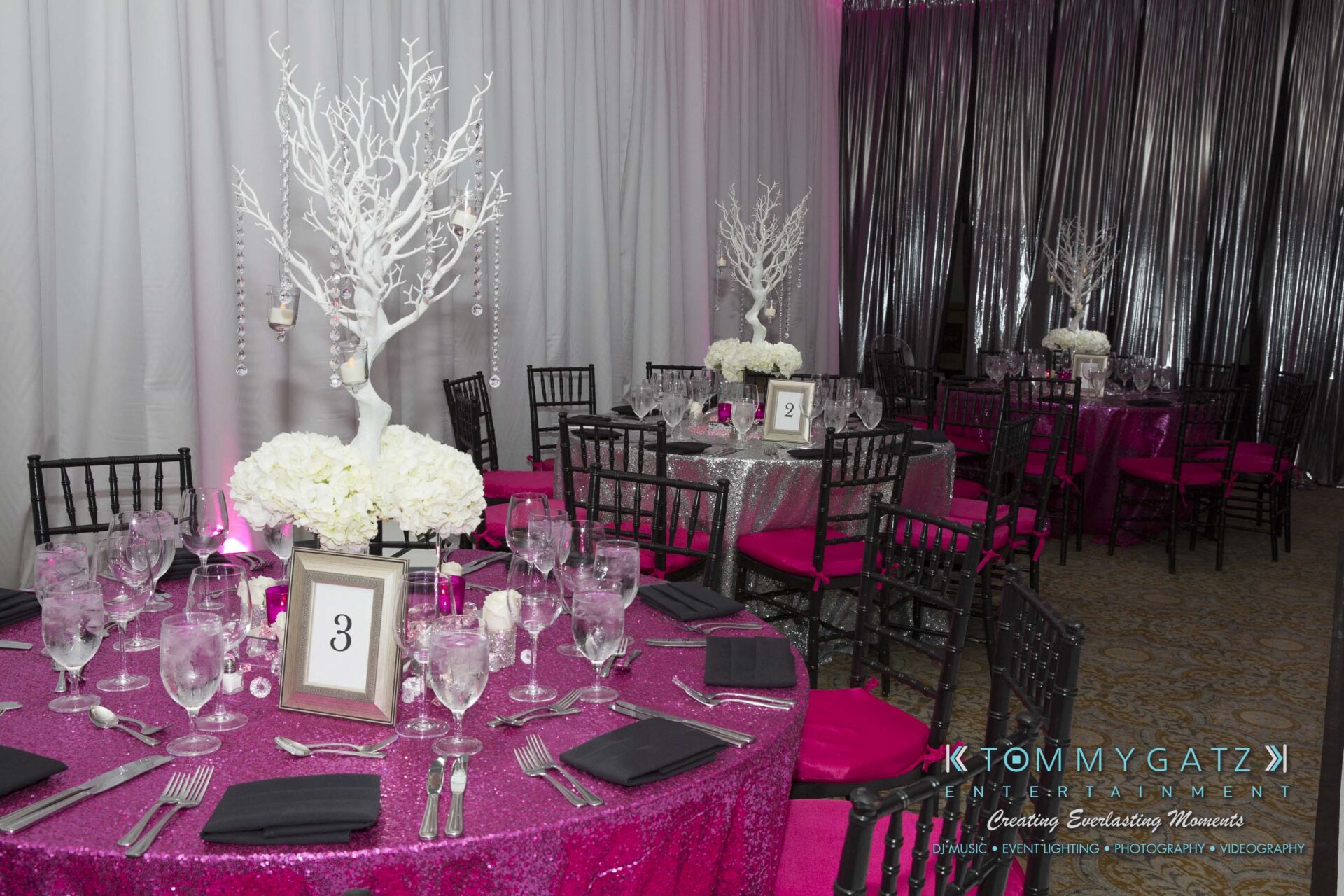 Dylans-Bat-Mitzvah-RECEPTION-Low-Res-WM-19 - TOMMY GATZ ENTERTAINMENT Elegant event setup with round tables covered in pink sequin tablecloths, black chairs, and silver centerpieces with white flowers. Numbered table signs are displayed.