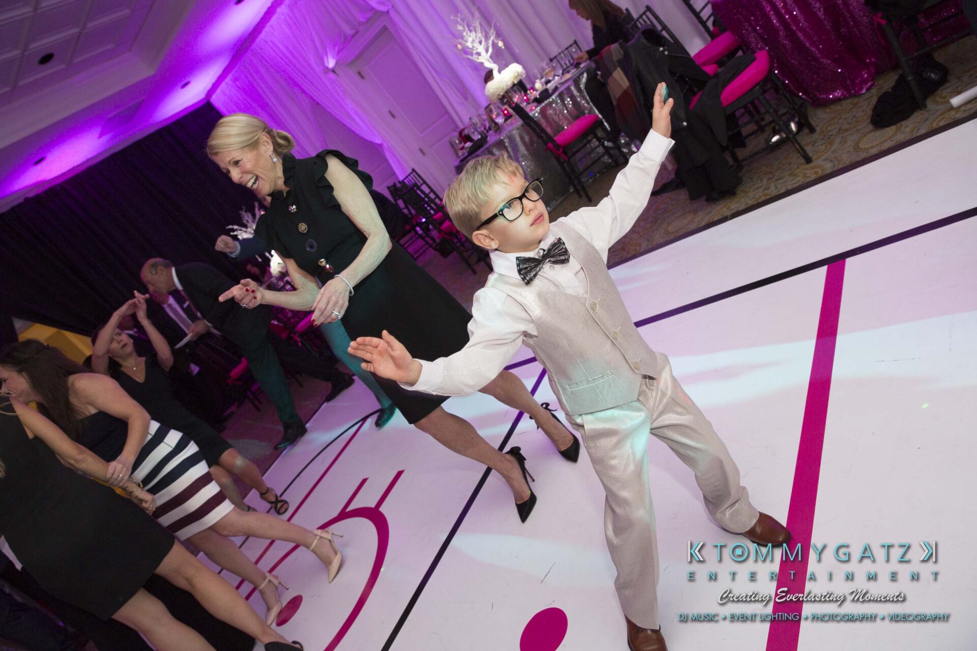 Dylans-Bat-Mitzvah-RECEPTION-Low-Res-WM-199 - TOMMY GATZ ENTERTAINMENT A young boy in a light-colored suit and glasses dances energetically on a dance floor, with people enjoying the event in the background.