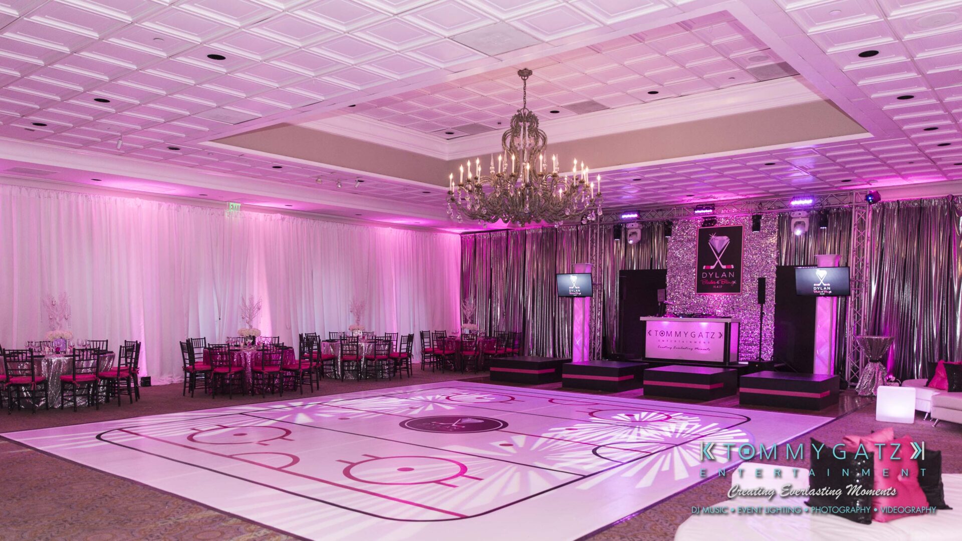 Dylans-Bat-Mitzvah-RECEPTION-Low-Res-WM-2 - TOMMY GATZ ENTERTAINMENT Lavish event venue with a pink-lit decor, featuring a dance floor resembling an ice rink, elegant drapes, and a chandelier. Tables and chairs are arranged around the floor.