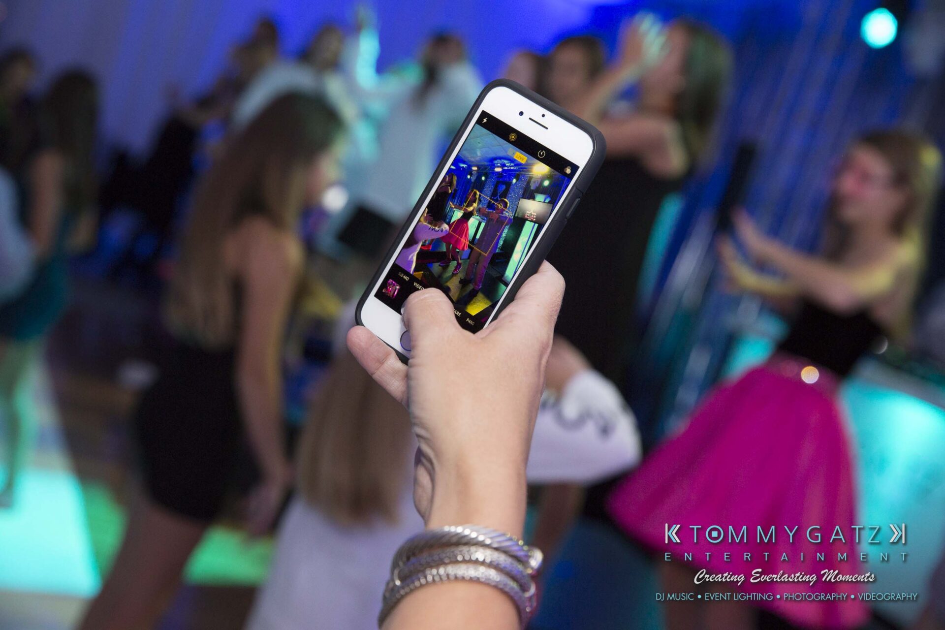Dylans-Bat-Mitzvah-RECEPTION-Low-Res-WM-200 - TOMMY GATZ ENTERTAINMENT Close-up of a person holding a smartphone, capturing a photo of people dancing at a lively event.