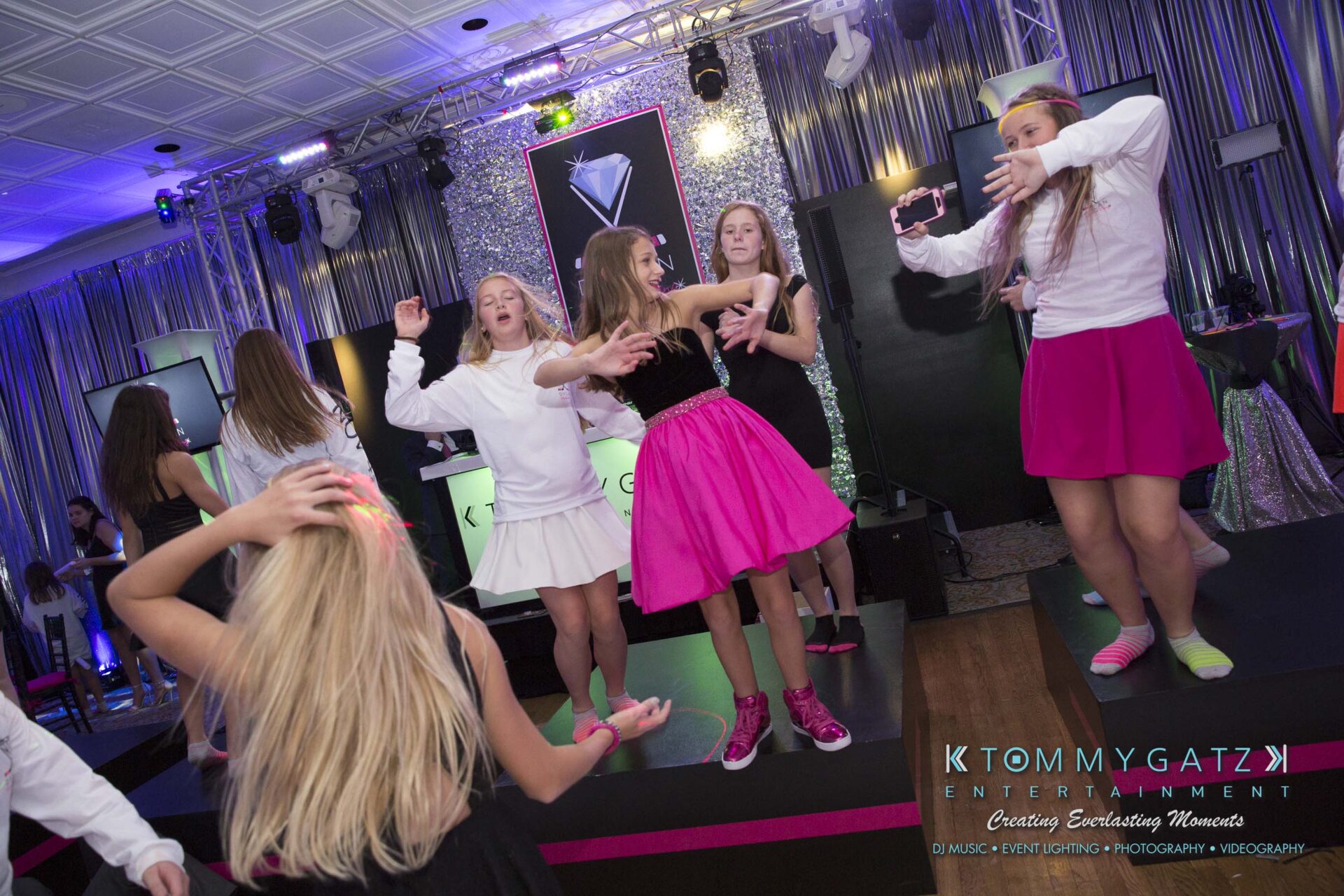 Dylans-Bat-Mitzvah-RECEPTION-Low-Res-WM-209 - TOMMY GATZ ENTERTAINMENT Group of young people dancing in a decorated indoor venue with a DJ booth and colorful lighting.