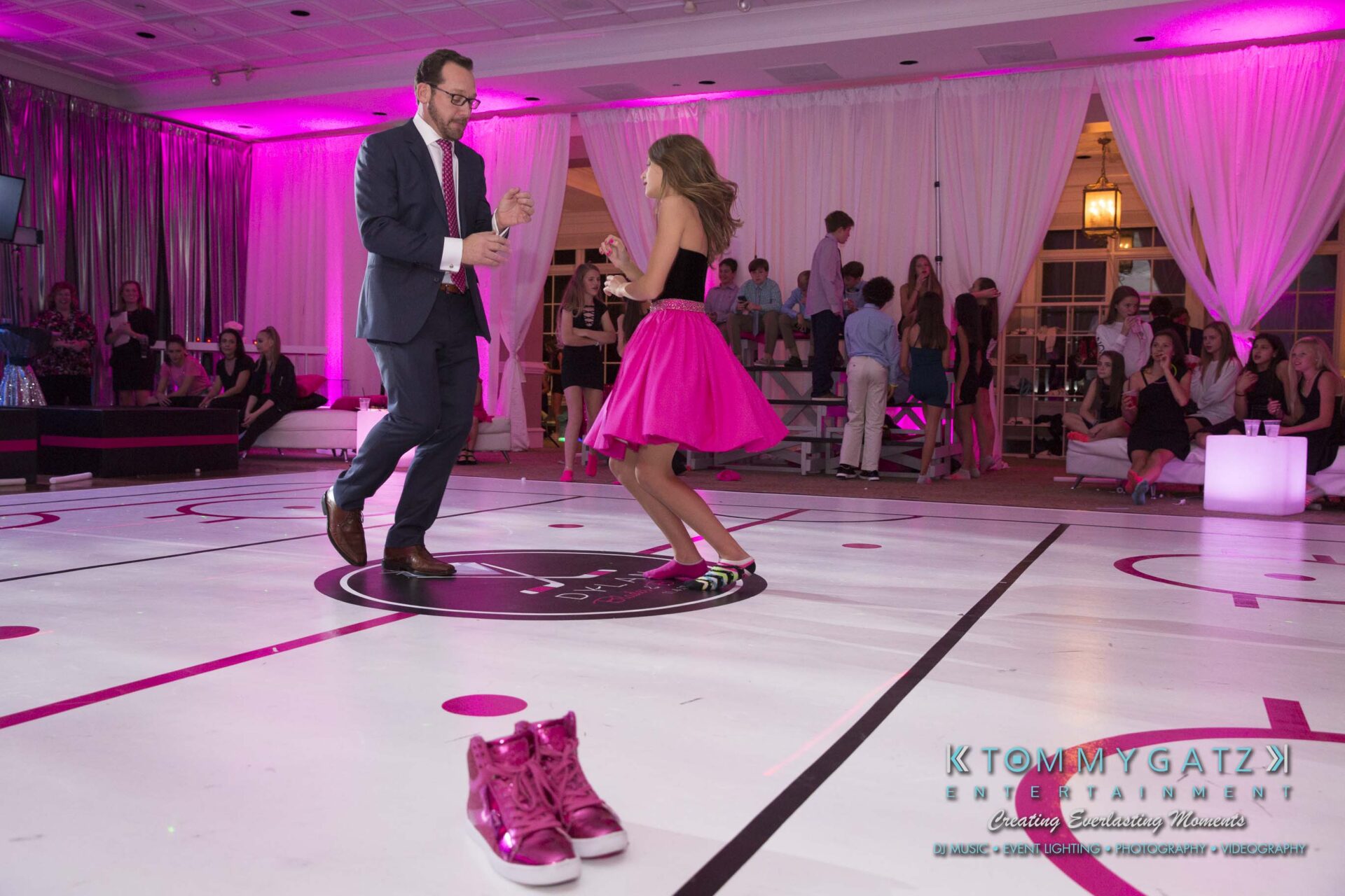 Dylans-Bat-Mitzvah-RECEPTION-Low-Res-WM-212 - TOMMY GATZ ENTERTAINMENT A man in a suit dances with a girl in a pink dress on a white dance floor. Pink shoes are on the floor nearby. The setting has pink and purple lighting.