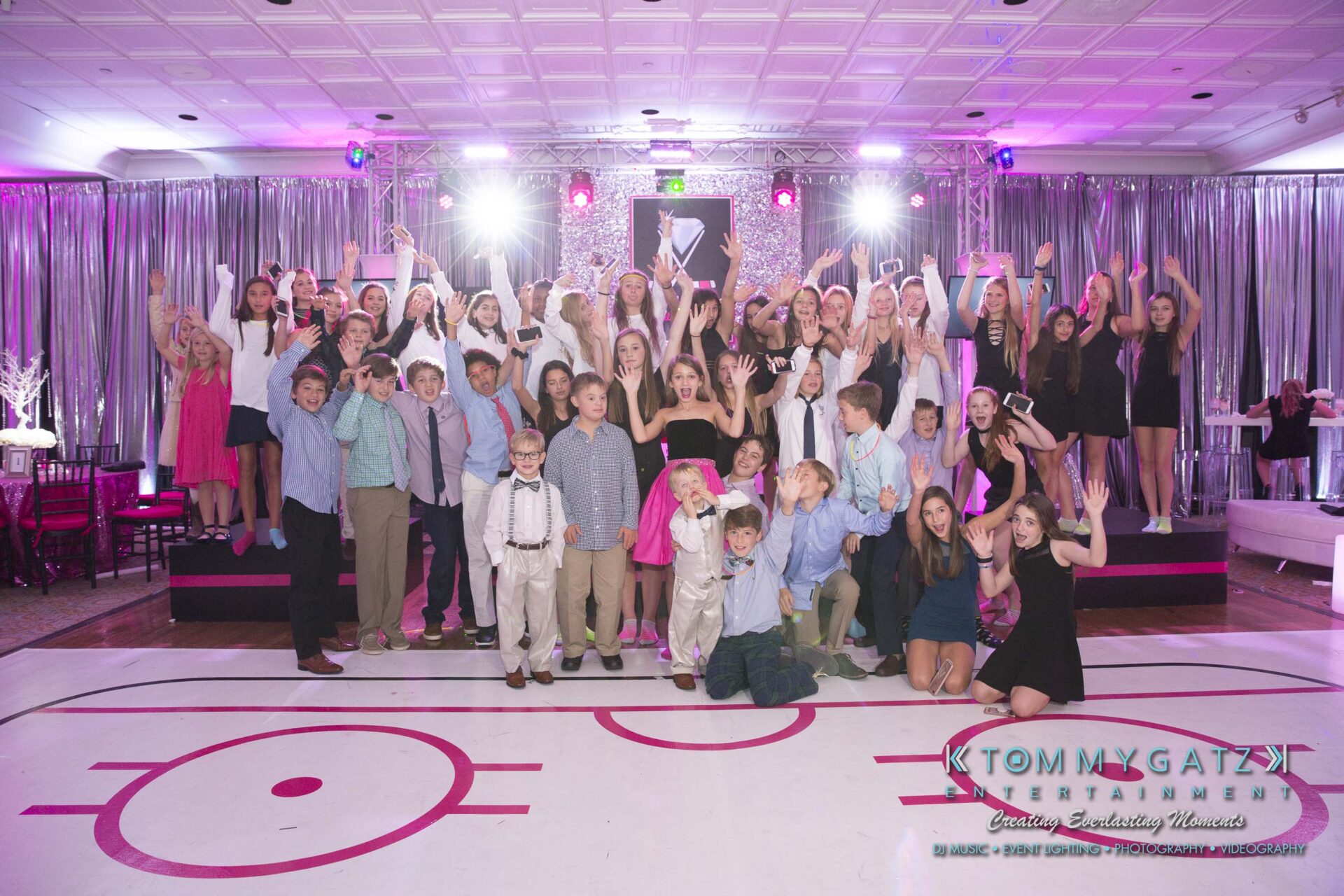 Dylans-Bat-Mitzvah-RECEPTION-Low-Res-WM-234 - TOMMY GATZ ENTERTAINMENT A large group of people, including children and adults, pose happily on a stage with arms raised. Bright lights and pink decorations are in the background.