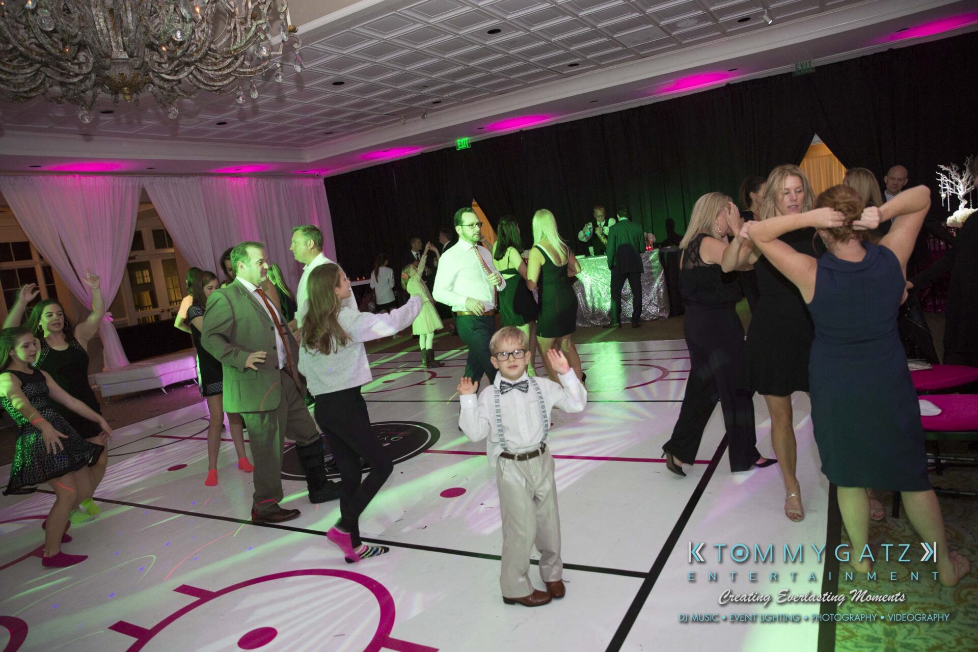 Dylans-Bat-Mitzvah-RECEPTION-Low-Res-WM-239 - TOMMY GATZ ENTERTAINMENT People dancing at a formal event with colorful lighting and decorations. A child stands in the foreground wearing glasses and a bow tie.