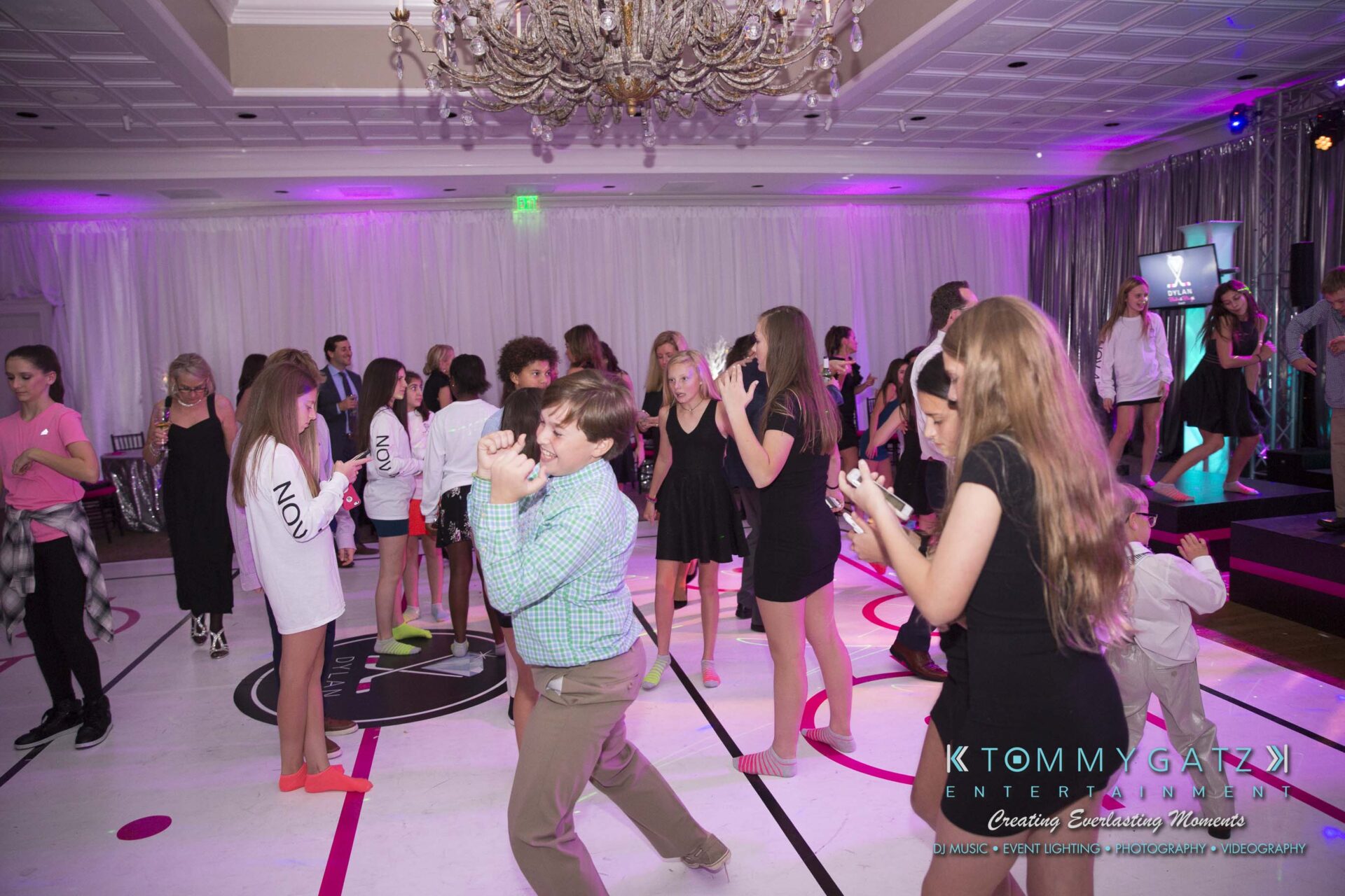 Dylans-Bat-Mitzvah-RECEPTION-Low-Res-WM-253-1 - TOMMY GATZ ENTERTAINMENT A group of people dancing and socializing at a lively event with colorful lighting and a decorative chandelier overhead.