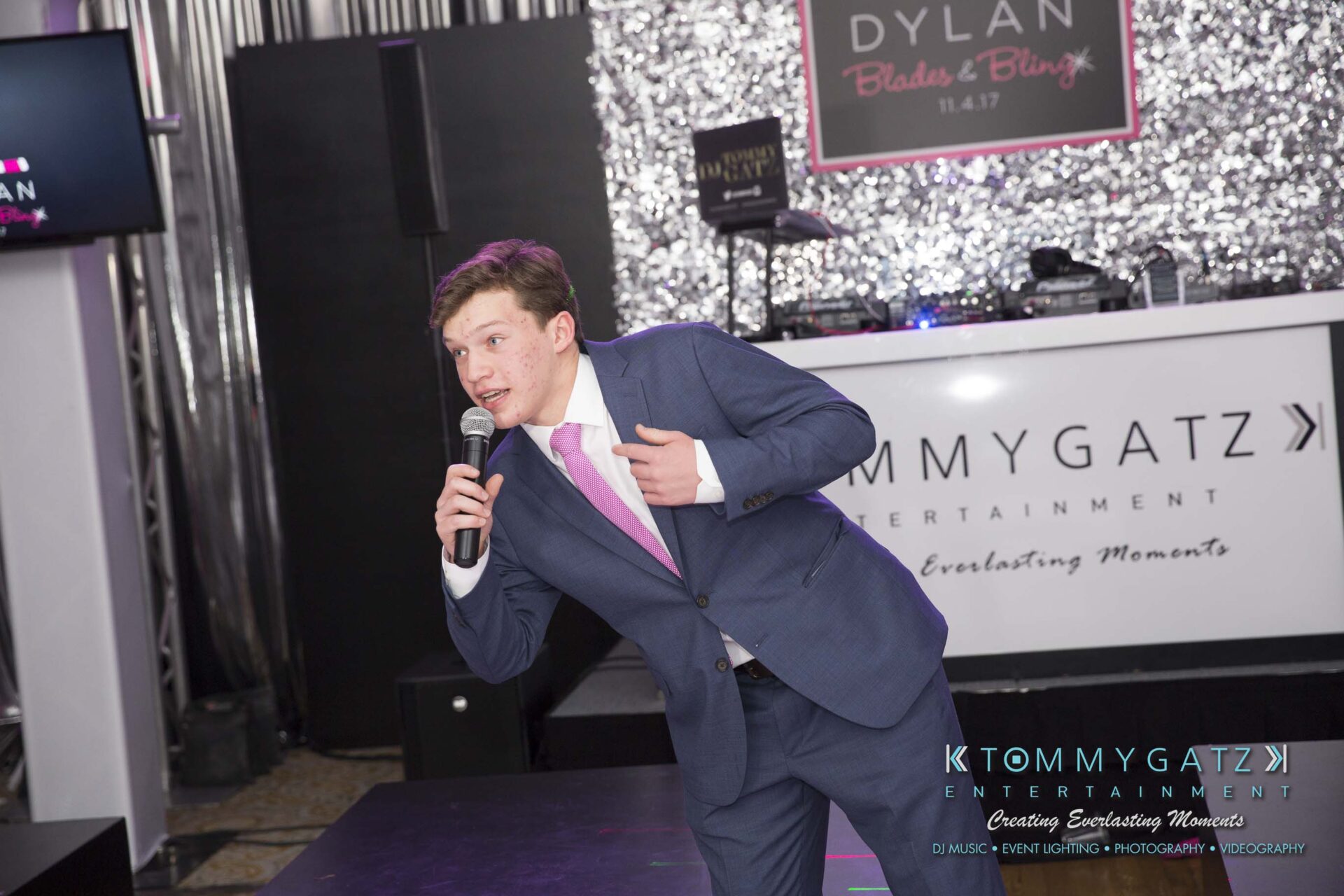 Dylans-Bat-Mitzvah-RECEPTION-Low-Res-WM-264 - TOMMY GATZ ENTERTAINMENT A man in a blue suit and pink tie holds a microphone, speaking on stage. A DJ booth with signage and glittery backdrop are behind him.