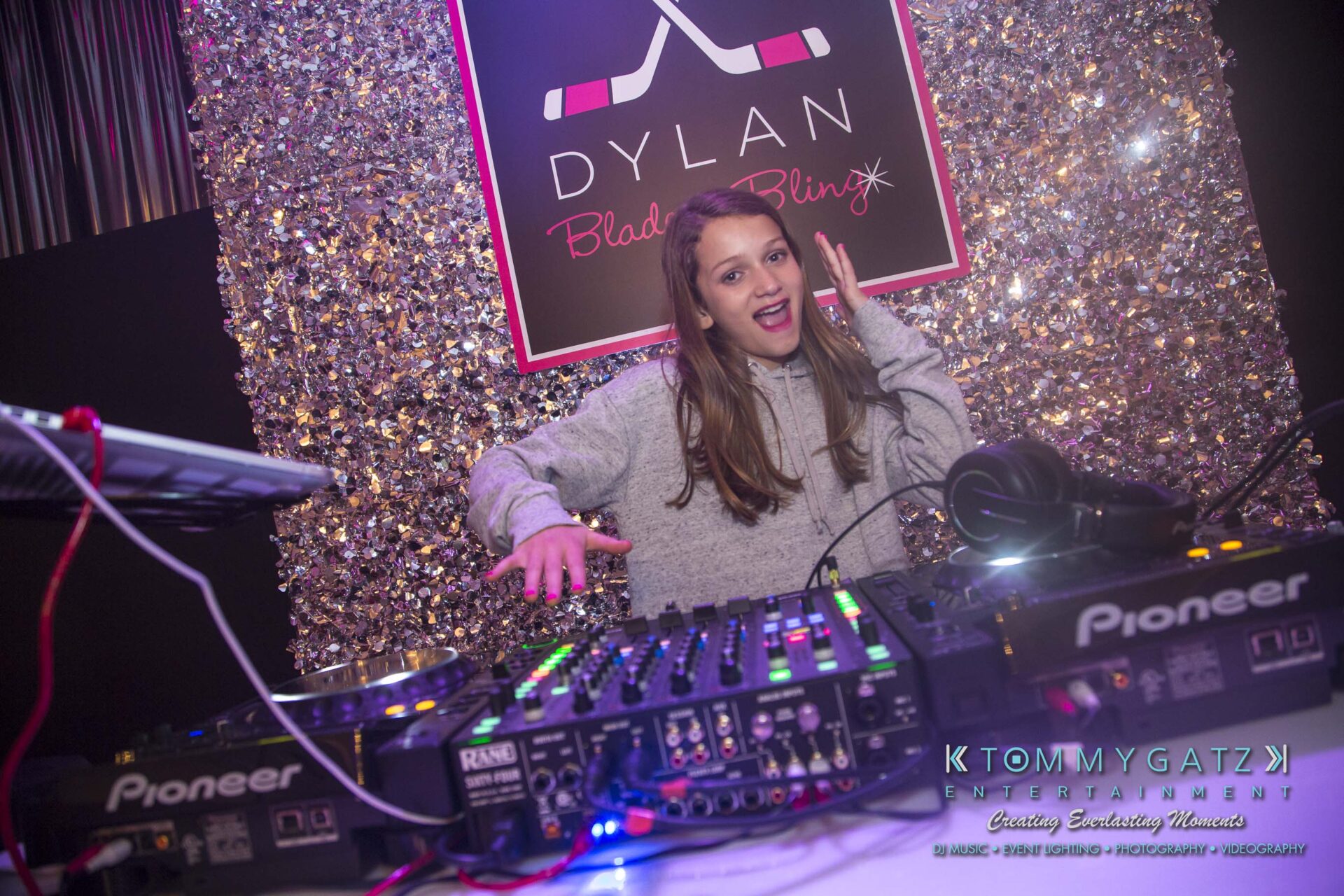 Dylans-Bat-Mitzvah-RECEPTION-Low-Res-WM-270 - TOMMY GATZ ENTERTAINMENT Person DJing at an event, standing behind a DJ console with colorful lights and a glittery backdrop.