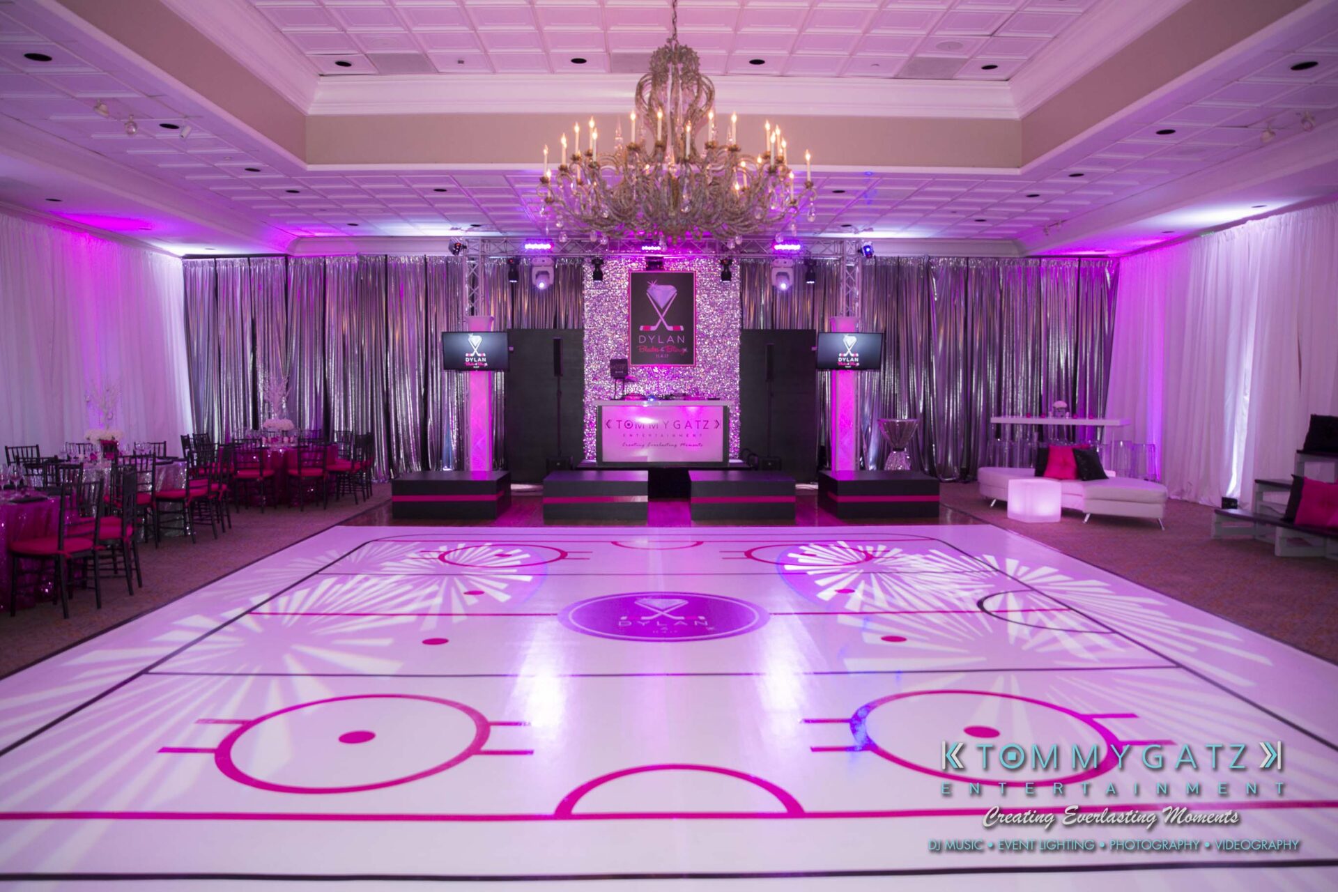 Dylans-Bat-Mitzvah-RECEPTION-Low-Res-WM-4 - TOMMY GATZ ENTERTAINMENT An event space with a hockey rink-themed floor, pink lighting, tables with black chairs, and a lit chandelier overhead.