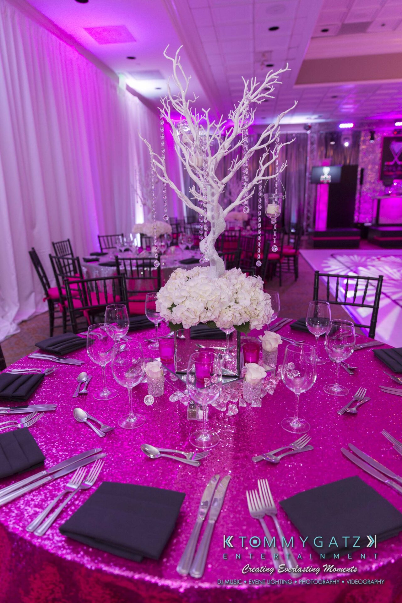 Dylans-Bat-Mitzvah-RECEPTION-Low-Res-WM-5 - TOMMY GATZ ENTERTAINMENT A table set for an event with black chairs, featuring a centerpiece of white flowers and a decorative white branch, on a pink sequin tablecloth.