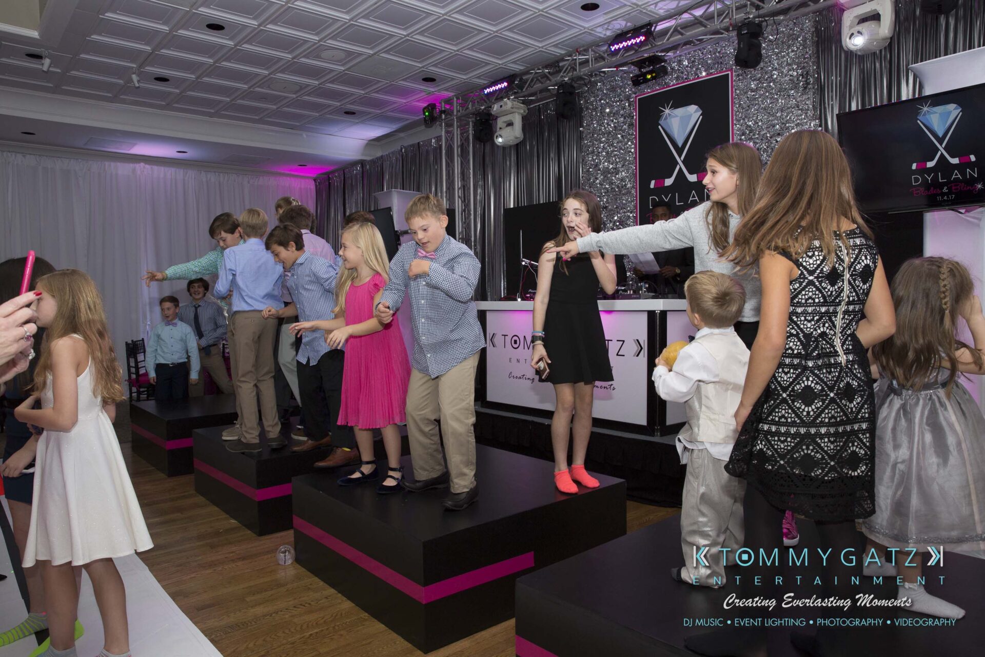 Dylans-Bat-Mitzvah-RECEPTION-Low-Res-WM-56 - TOMMY GATZ ENTERTAINMENT Group of children dancing on a stage at an event with pink and purple lighting. DJ booth and banners in the background.