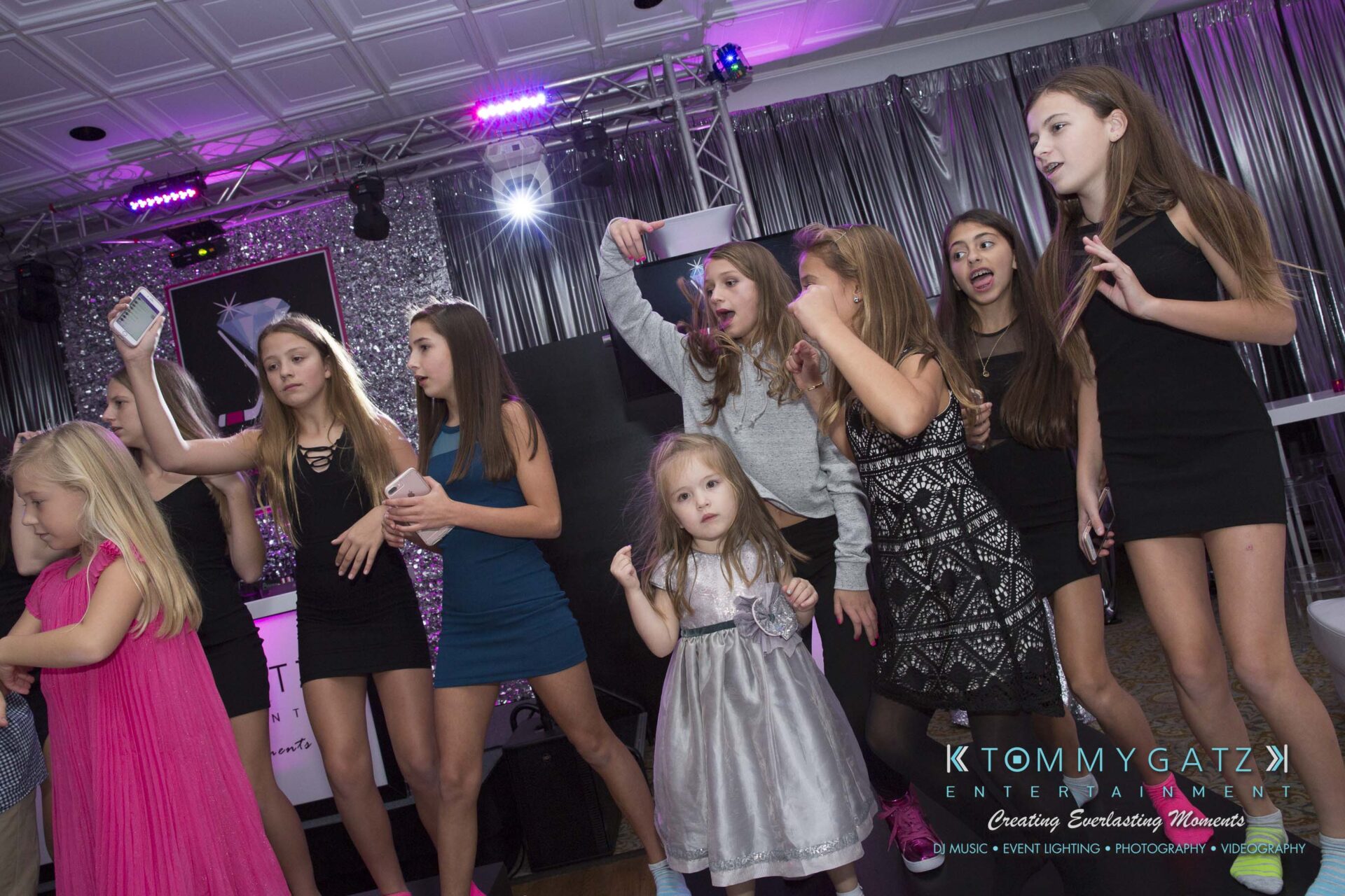 Dylans-Bat-Mitzvah-RECEPTION-Low-Res-WM-67 - TOMMY GATZ ENTERTAINMENT A group of children and teenagers in a party setting, some holding phones and striking poses, in a room with shiny decor and a DJ booth in the background.