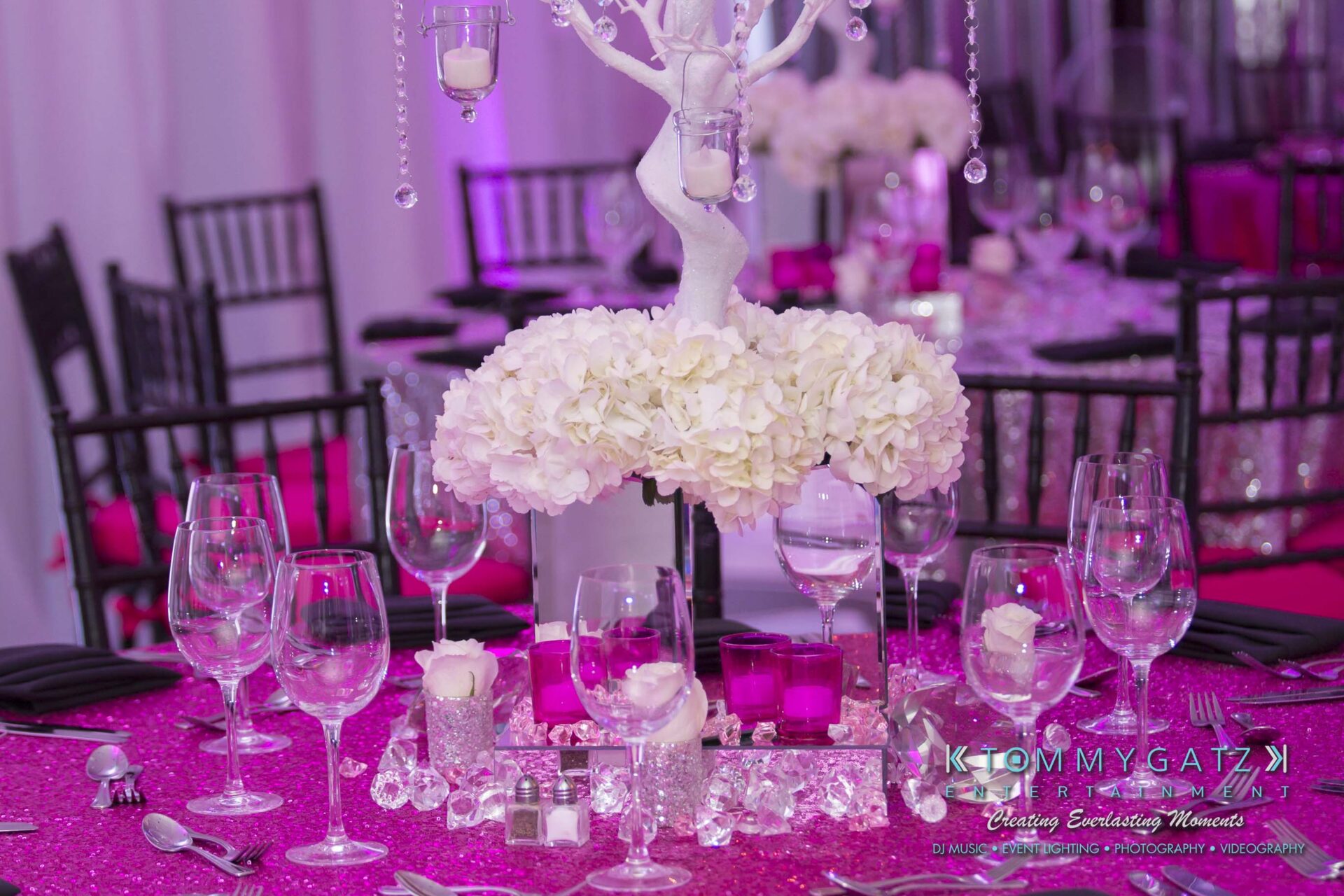 Dylans-Bat-Mitzvah-RECEPTION-Low-Res-WM-8 - TOMMY GATZ ENTERTAINMENT A table set for an event with a centerpiece of white flowers and candles, surrounded by wine glasses and covered in a pink sequin tablecloth.