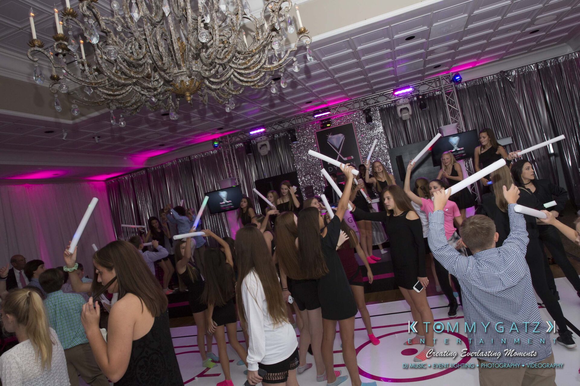 Dylans-Bat-Mitzvah-RECEPTION-Low-Res-WM-84 - TOMMY GATZ ENTERTAINMENT People dancing in an event hall with a chandelier, holding light sticks, and a DJ booth in the background.