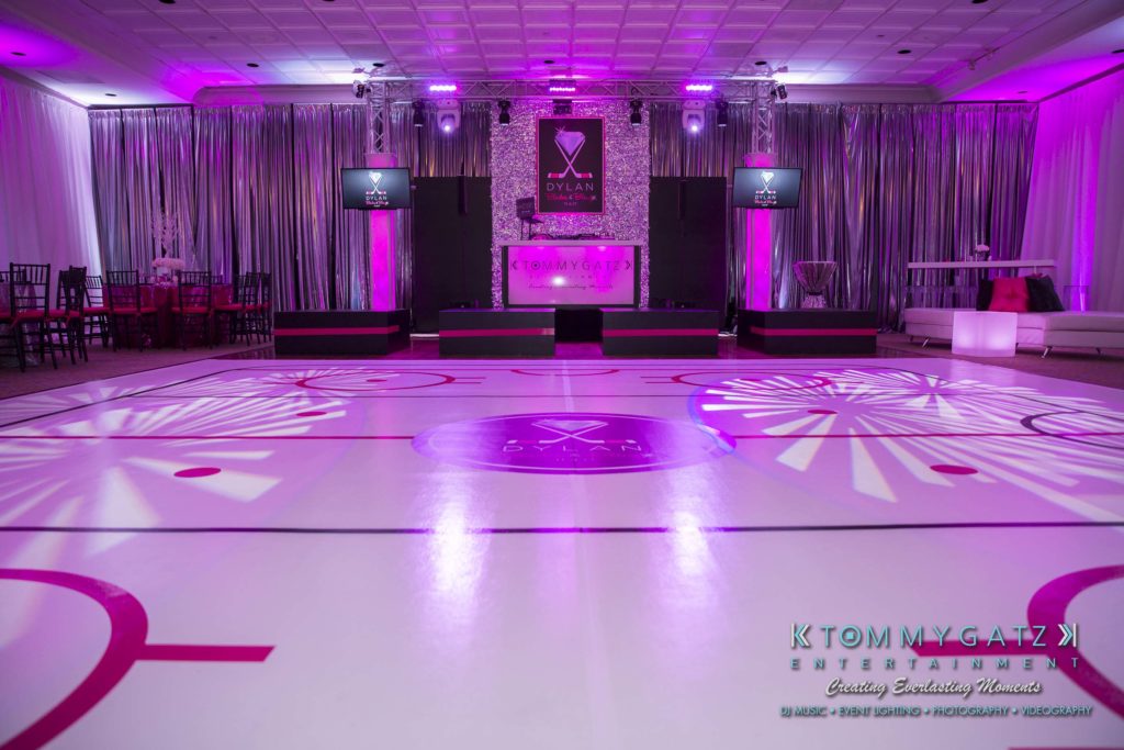 Dylans-Bat-Mitzvah-RECEPTION-Low-Res-WM-9-1024x683 - TOMMY GATZ ENTERTAINMENT A party room with purple lighting and a dance floor resembling a hockey rink, featuring DJ equipment on a stage.
