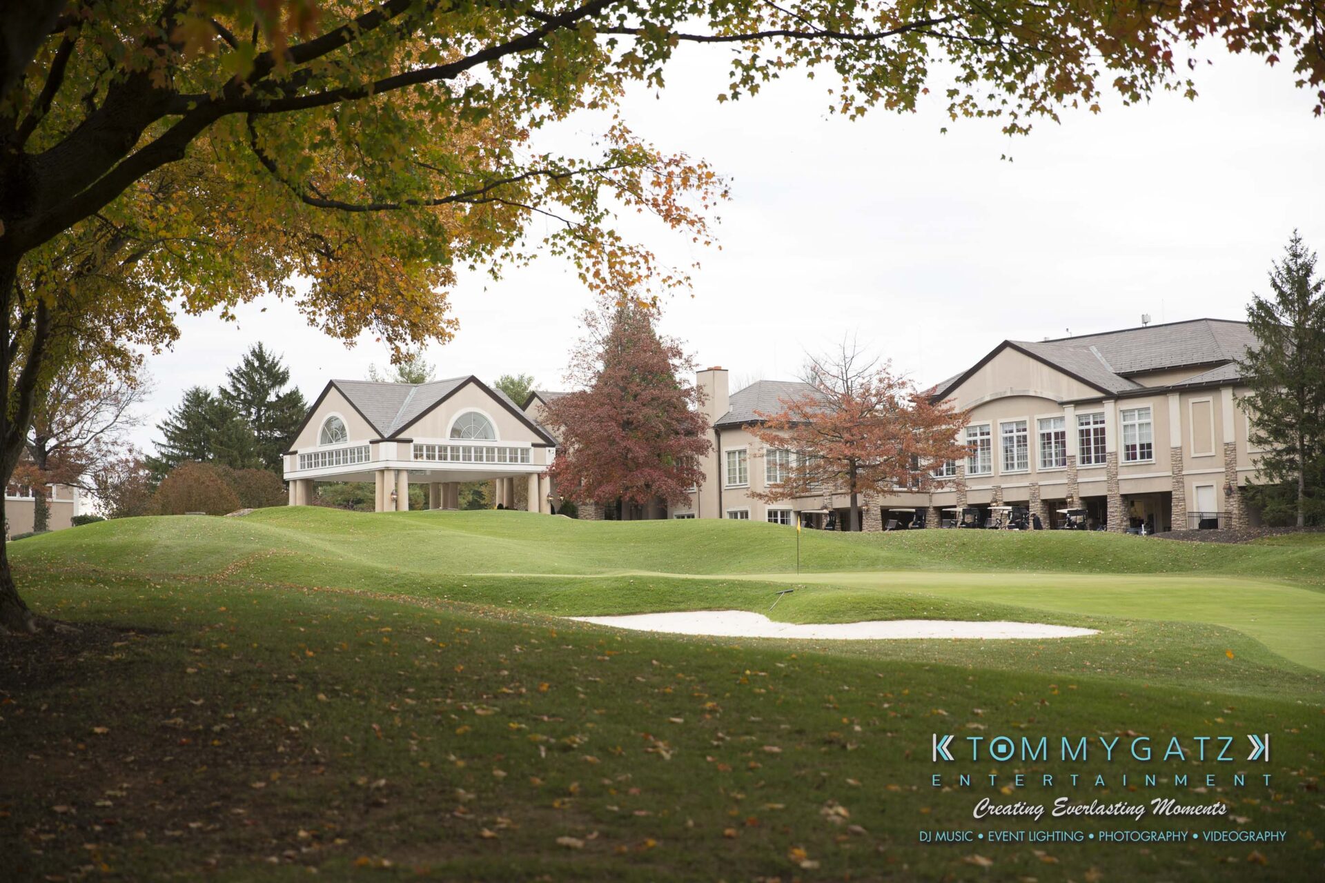 Dylans-Bat-Mitzvah-SOCIAL-HOUR-Low-Res-WM-2 - TOMMY GATZ ENTERTAINMENT A golf course with a sand trap in the foreground, surrounded by trees with autumn leaves. In the background, there are large buildings with expansive windows.