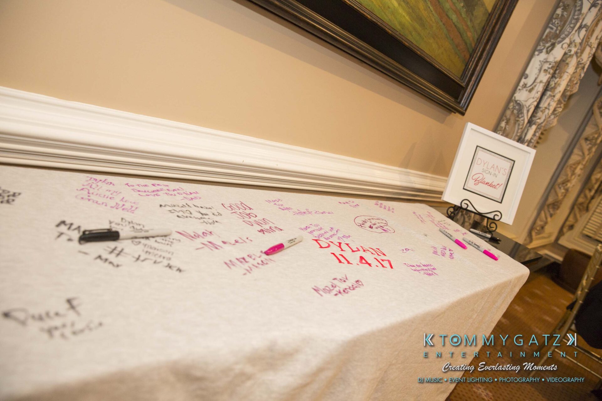 Dylans-Bat-Mitzvah-SOCIAL-HOUR-Low-Res-WM-34 - TOMMY GATZ ENTERTAINMENT Table with a tablecloth featuring handwritten messages in various colors, a black marker on top, a framed sign in the background, and decorative curtains nearby.