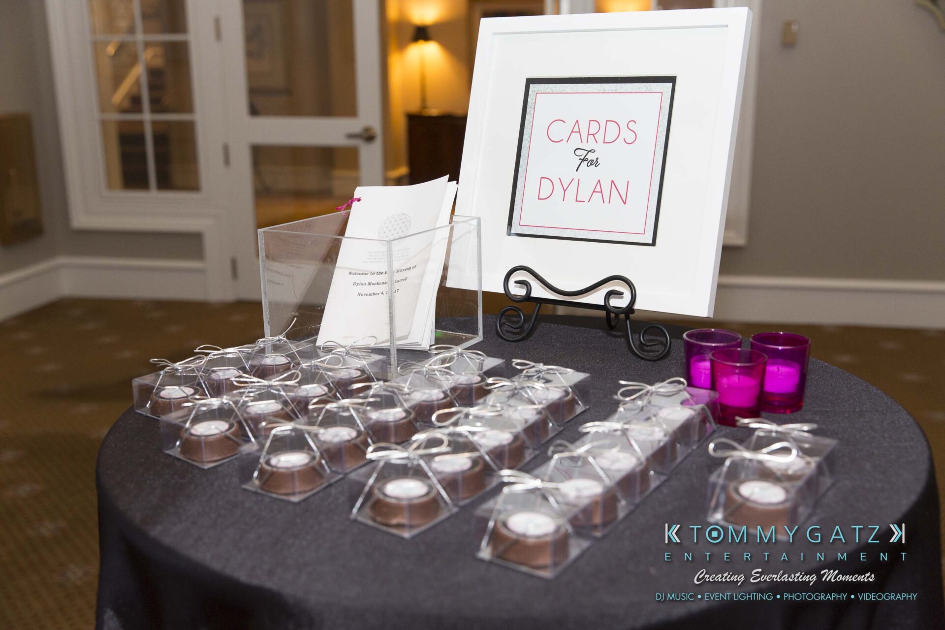 Dylans-Bat-Mitzvah-SOCIAL-HOUR-Low-Res-WM-68 - TOMMY GATZ ENTERTAINMENT A table with small gift boxes, a "Cards for Dylan" sign, and two purple candles displayed in a room.